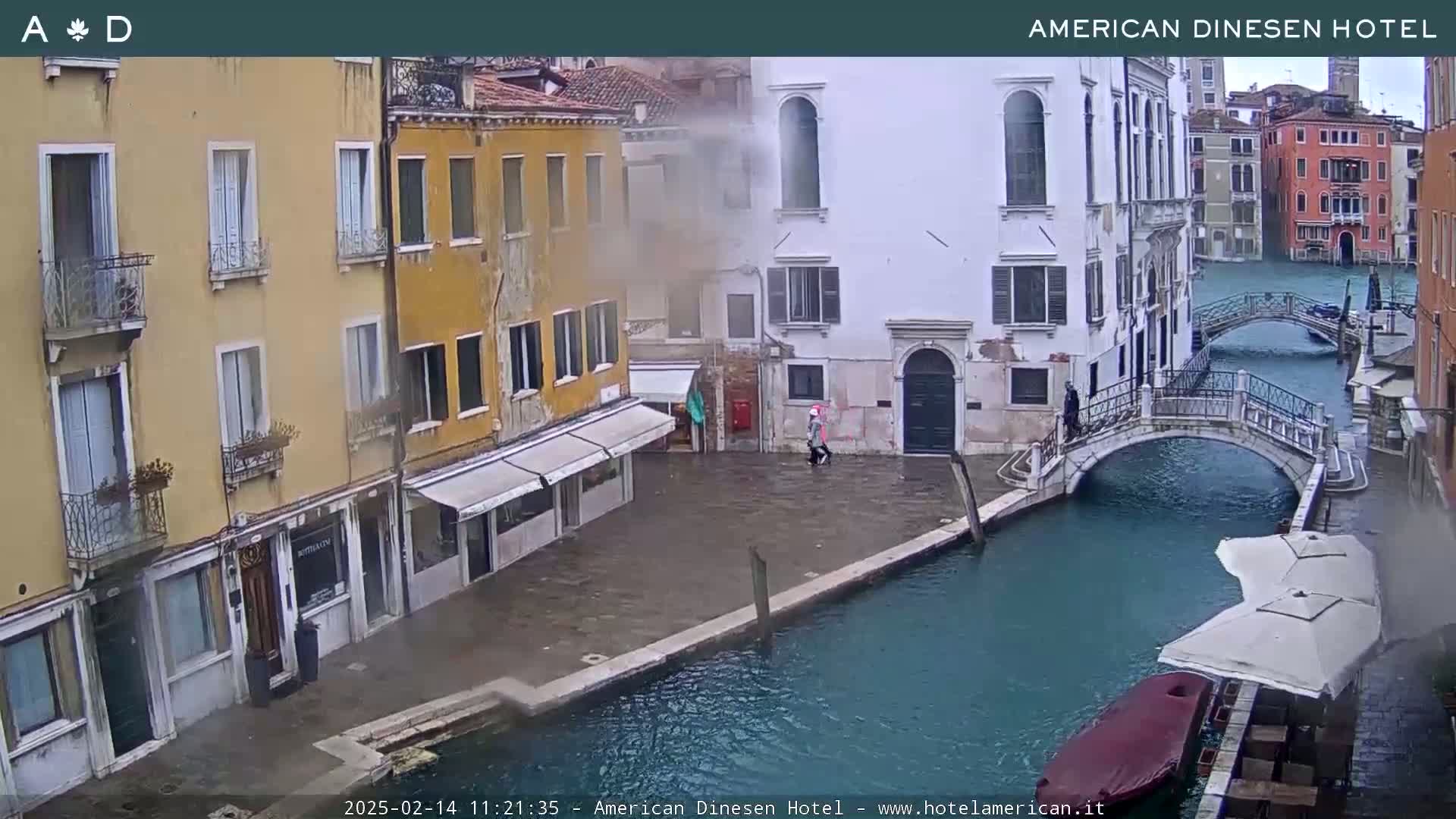 A view of a canal in Venice, Italy, on an overcast day, shows several buildings lining the waterway, with a small bridge and a few pedestrians visible.