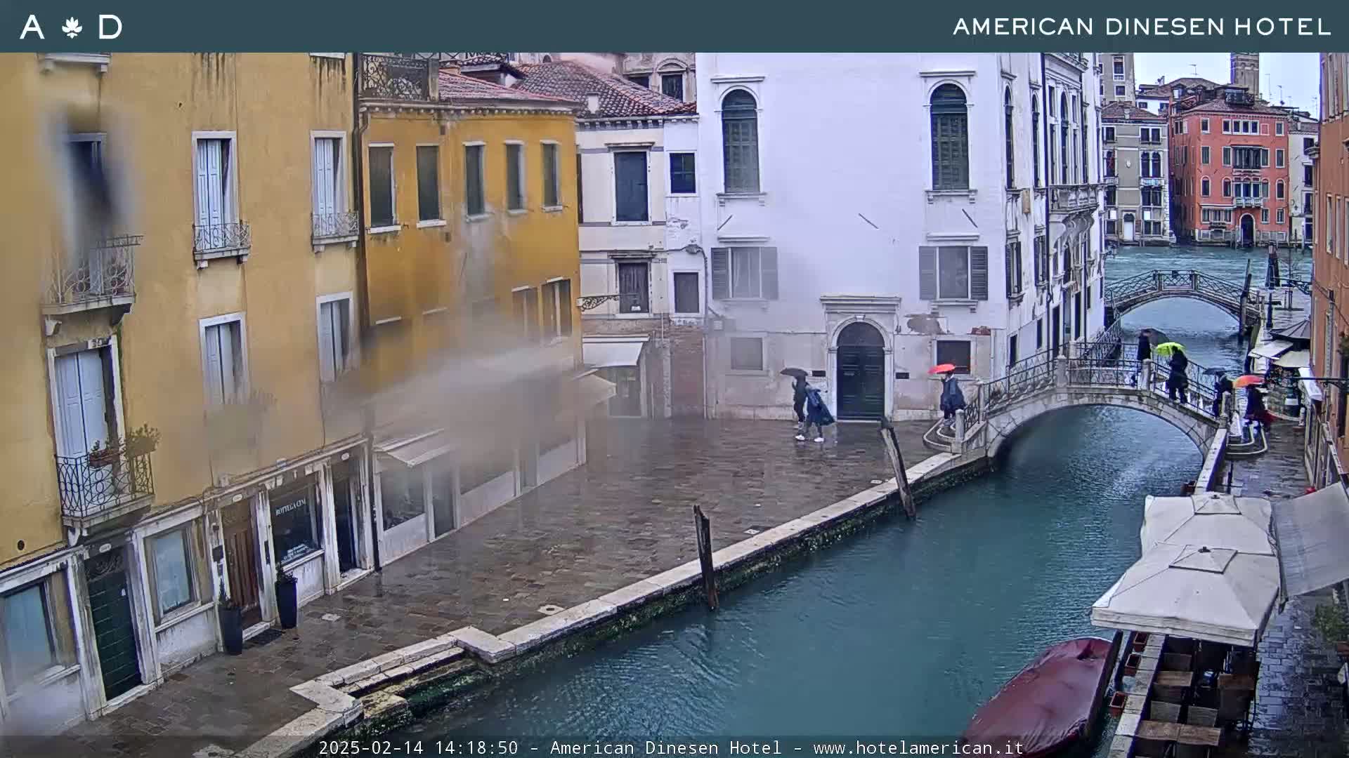 A rain-soaked Venetian canal scene shows several people with umbrellas walking across a bridge, alongside colorful buildings and a boat with patio umbrellas.
