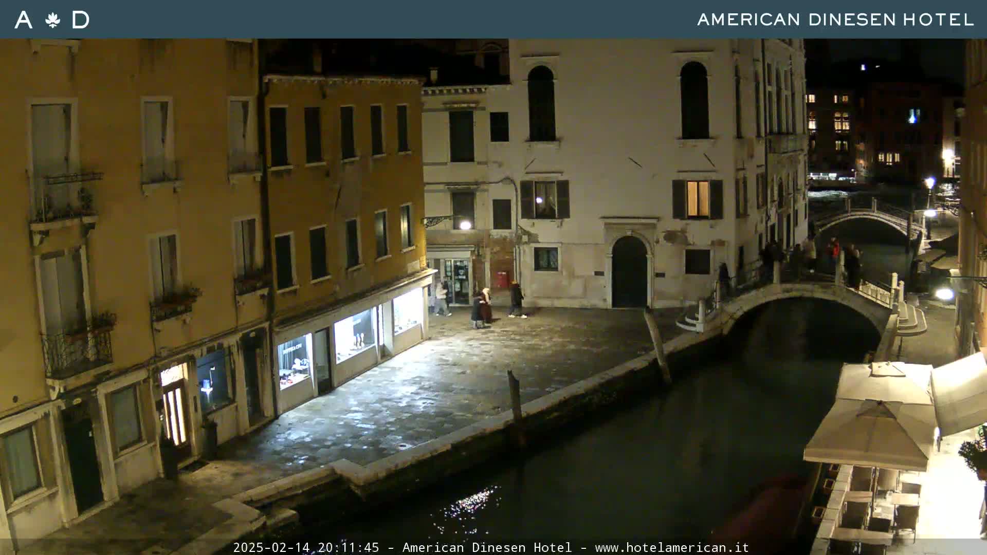 A nighttime view of a canal in Venice, showing several buildings, a bridge, and a few people gathered in a small plaza.