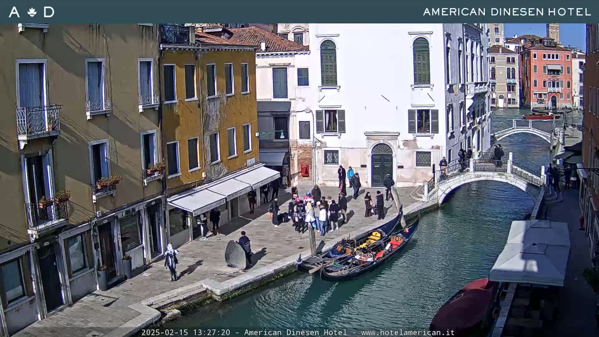 A sunny view of a Venetian canal with gondolas, pedestrians on a bridge and along the water's edge, and buildings lining the waterway.