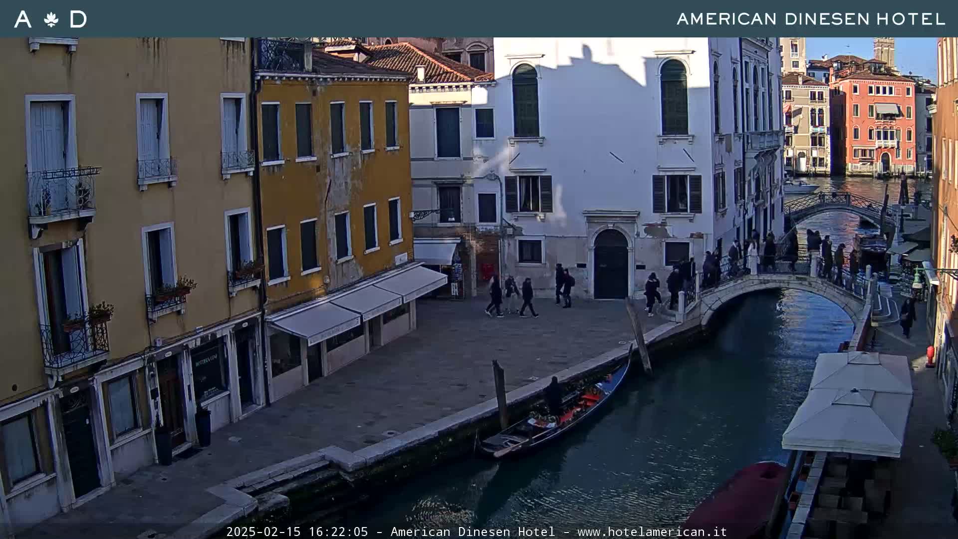 A Venetian canal scene on a sunny day shows pedestrians on a bridge crossing over the canal, gondolas in the water, and several buildings lining the canal's edge.