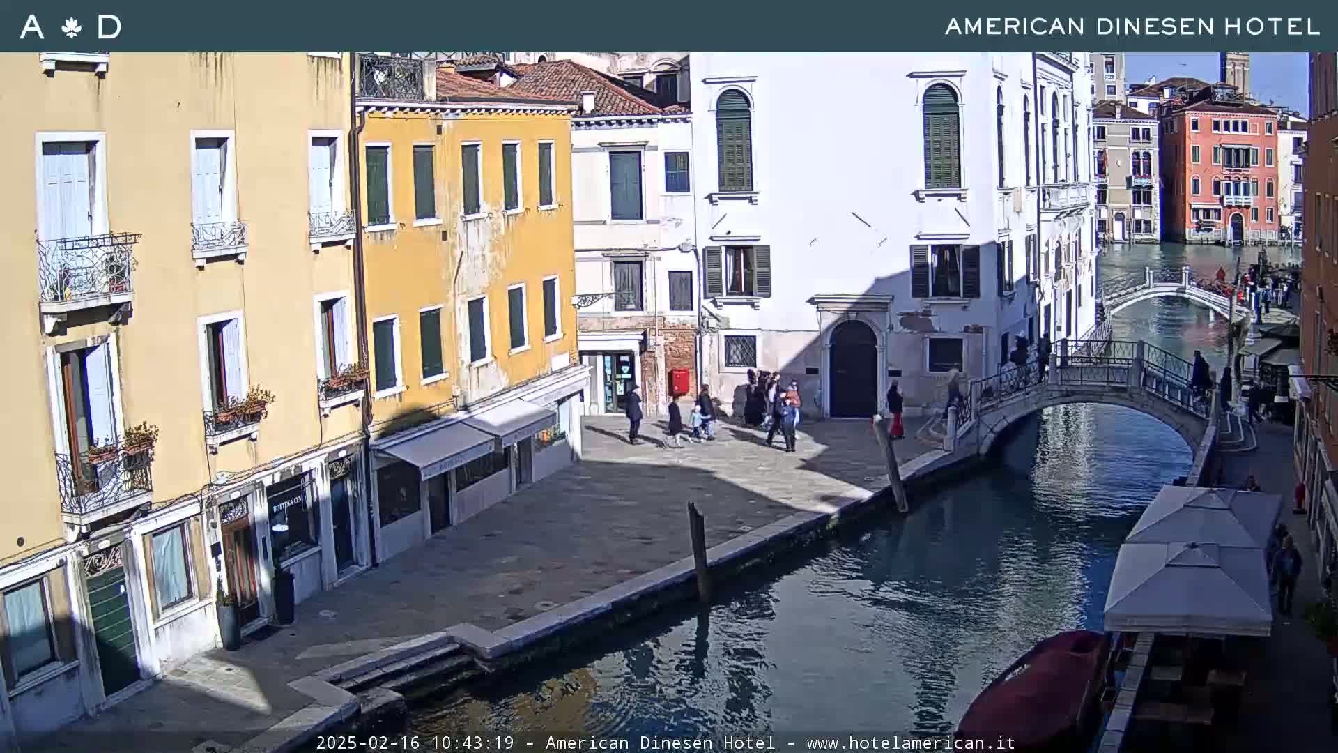 A sunny day in Venice shows a canal with a bridge, several buildings lining the waterway, and pedestrians walking along the canal bank.