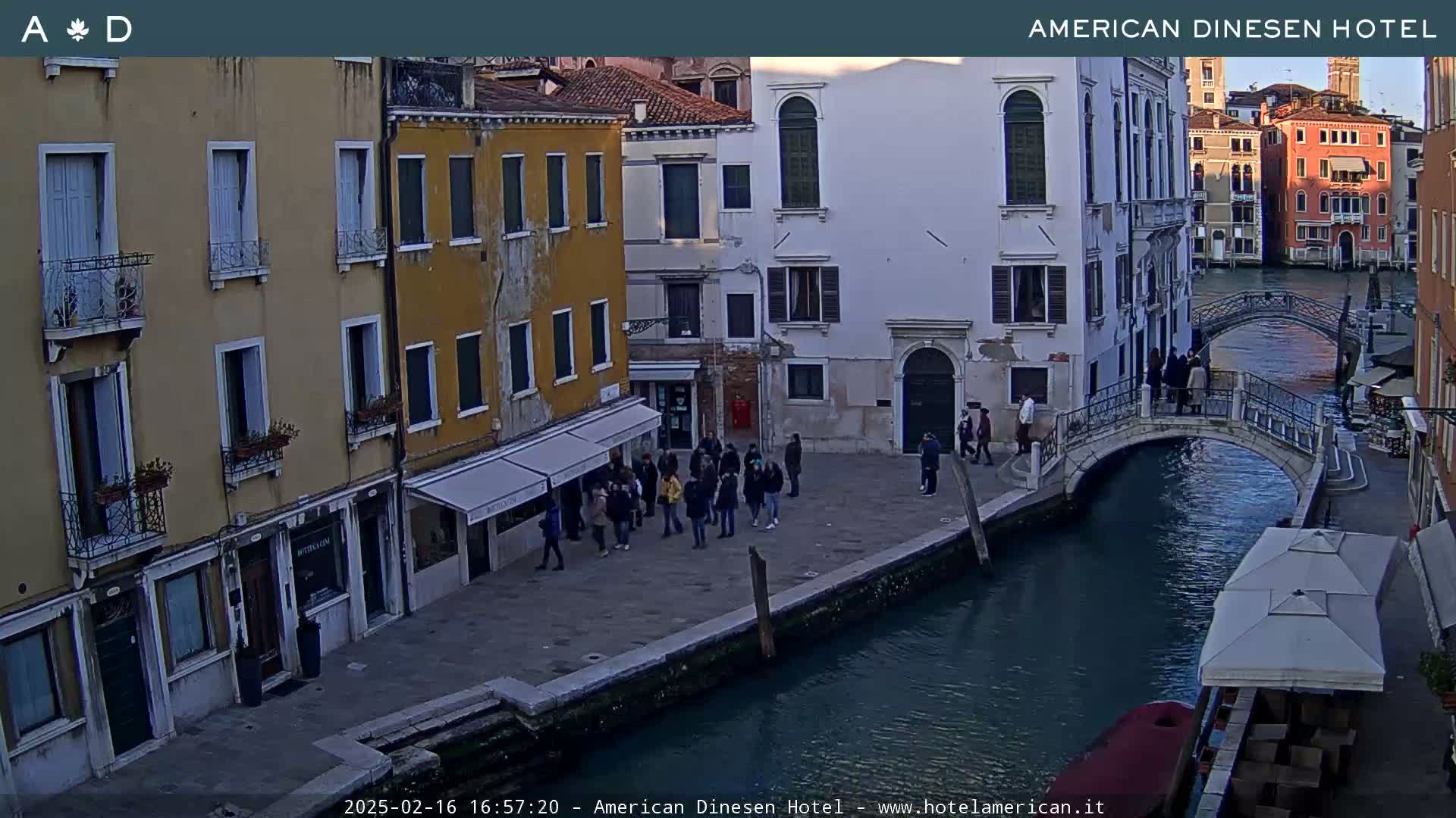 A view of a canal in Venice, Italy, shows several people walking on a bridge and near buildings along the waterfront, under a dusky sky.