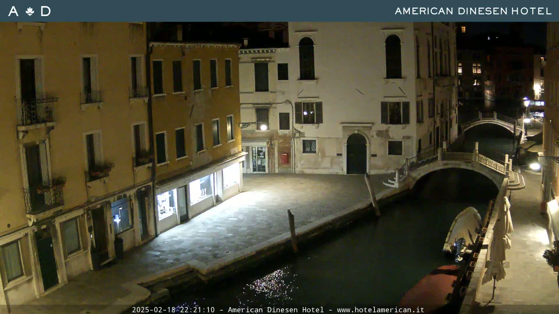 A nighttime view of a canal in Venice, Italy, shows several buildings lining the waterway, with a small boat moored alongside one of the bridges.