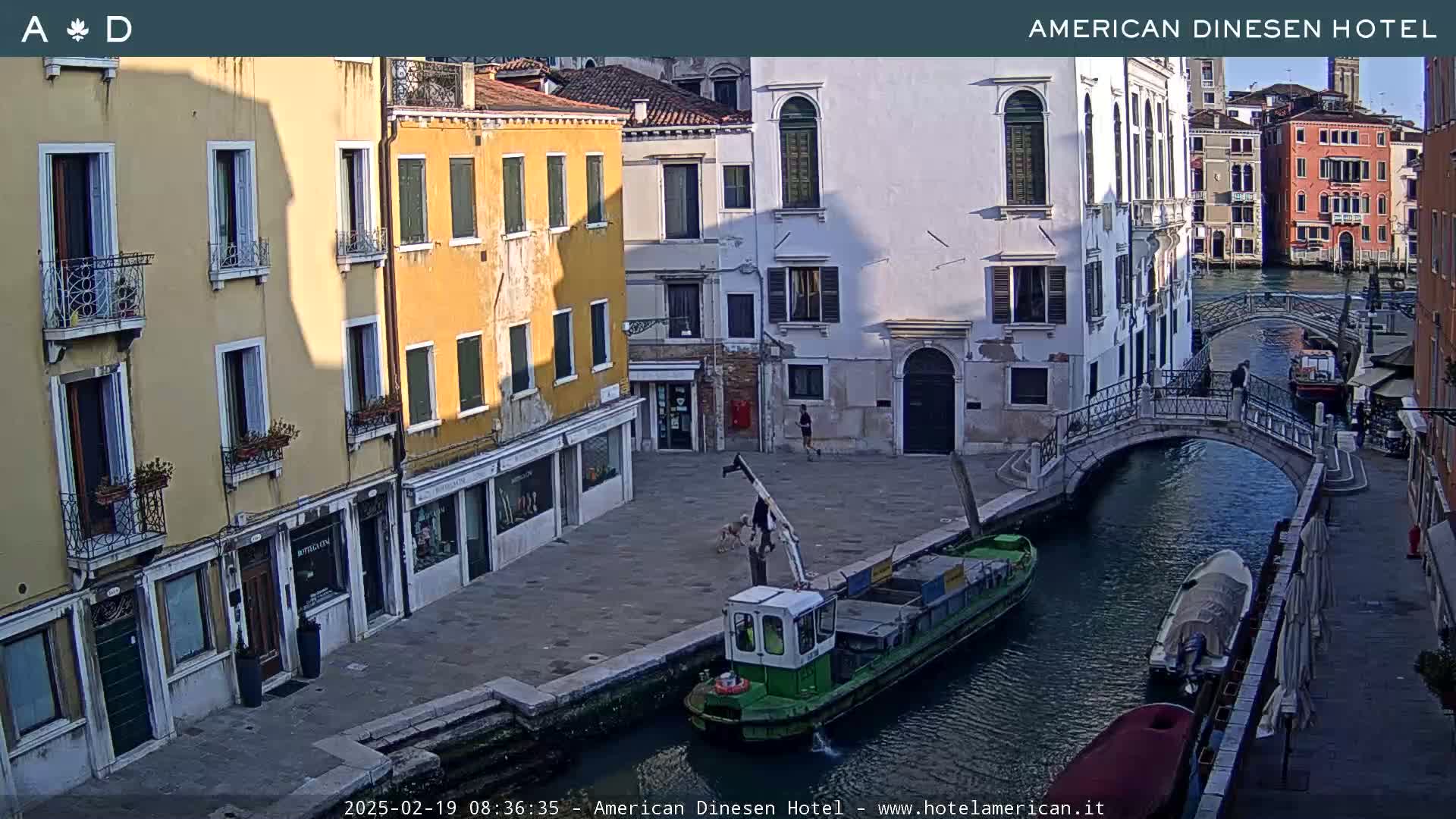 A sunny day in Venice shows a canal with a small green barge, a pedestrian bridge, and colorful buildings lining the waterway.