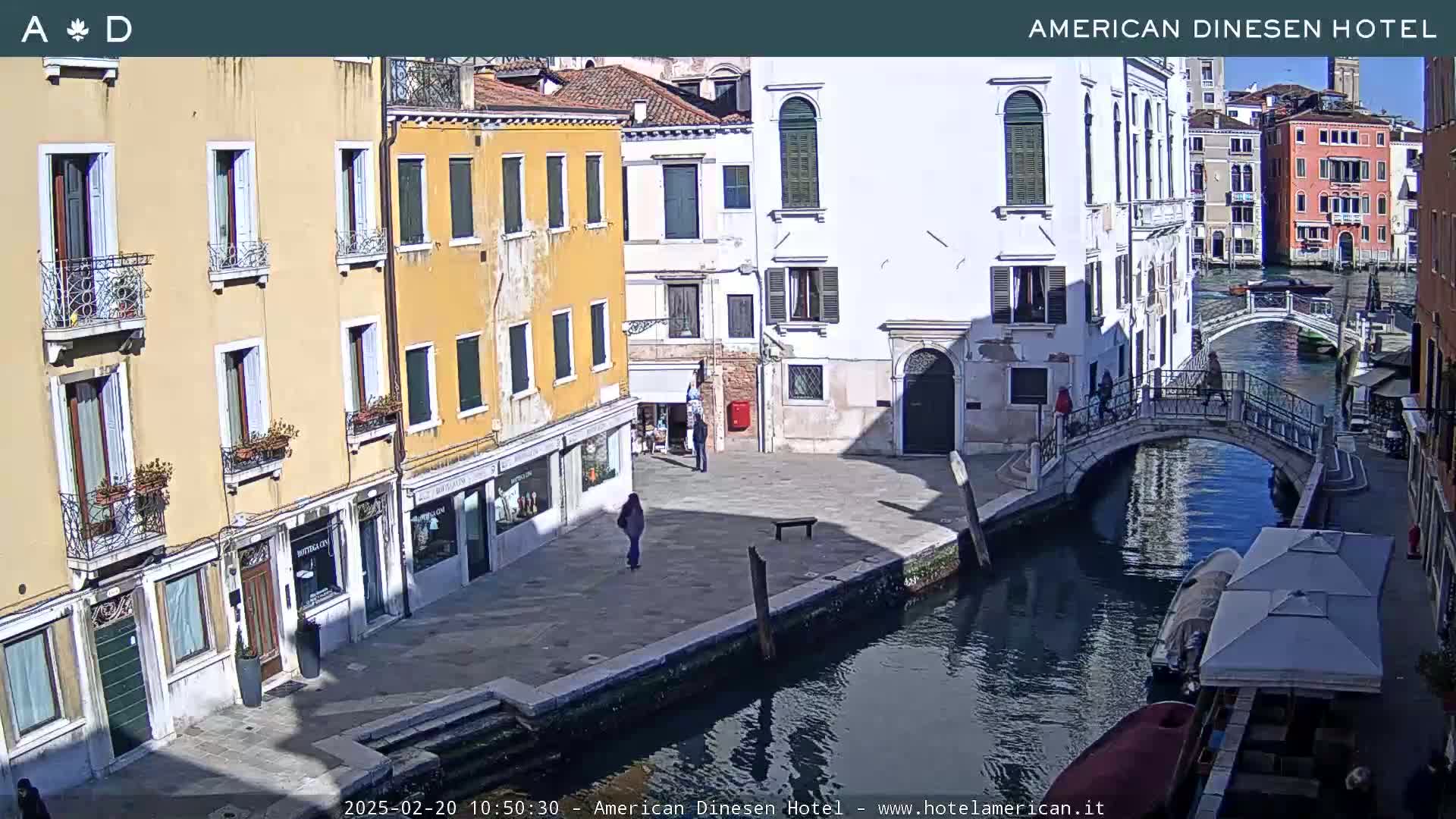 A sunny day in Venice shows a canal with a bridge, lined with closely-packed buildings, and a few pedestrians.