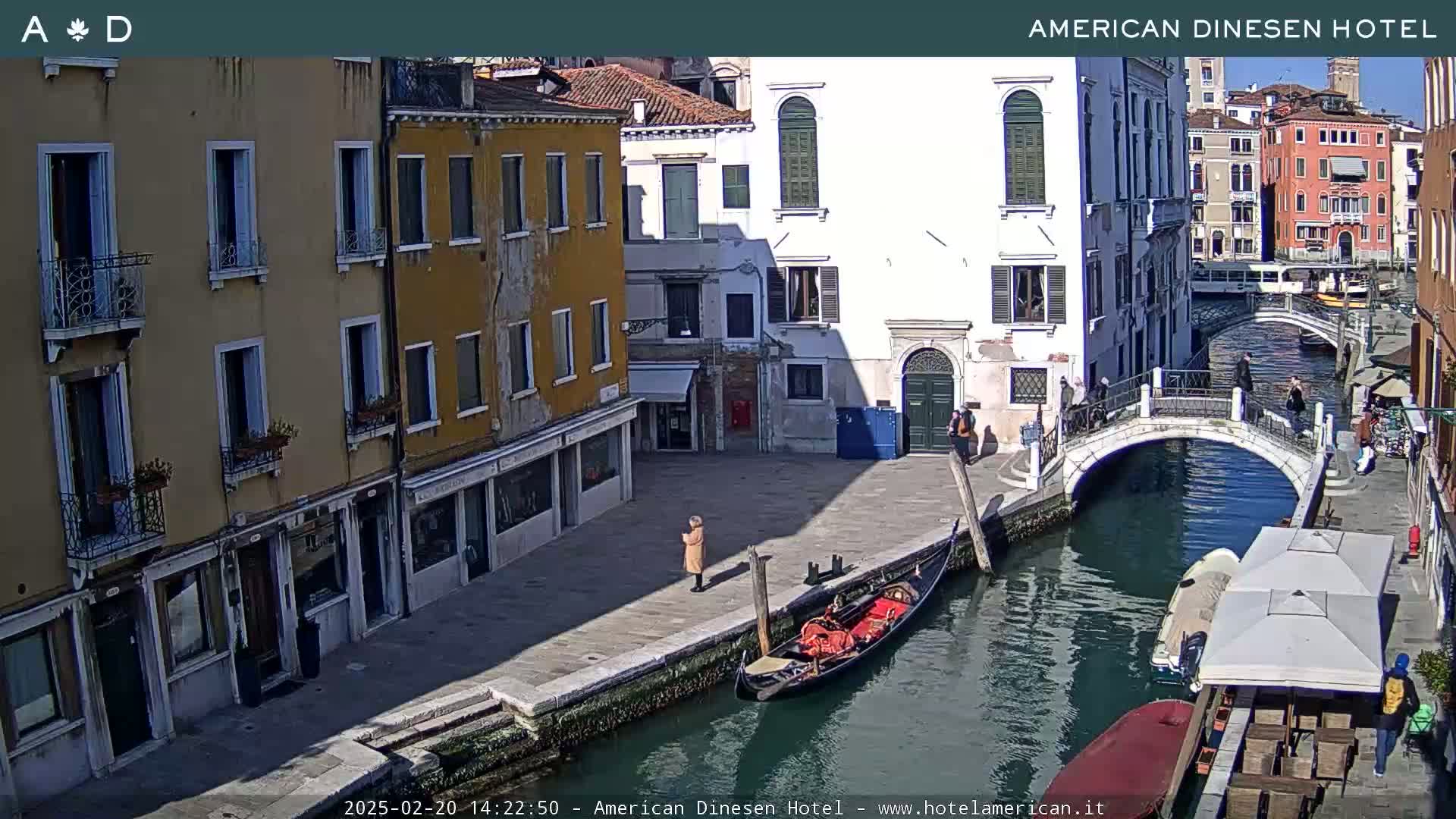 A sunny day in Venice shows a canal with a gondola, a pedestrian bridge, and buildings lining the waterway.