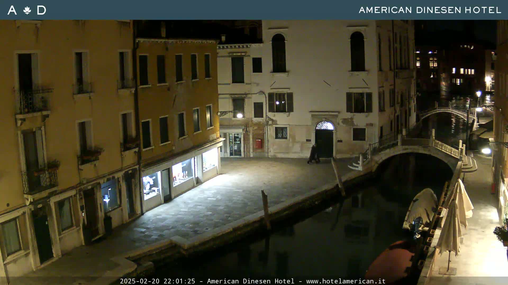 A nighttime view of a canal in Venice, showing buildings lining the waterway and a small bridge, under clear skies.