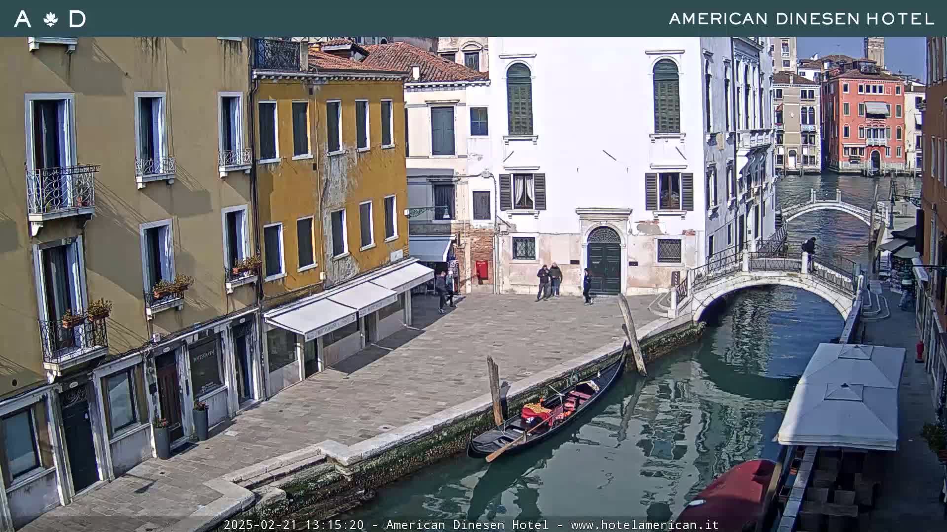 A sunny day in Venice shows a canal with a gondola, a small bridge, and several buildings lining the waterway.