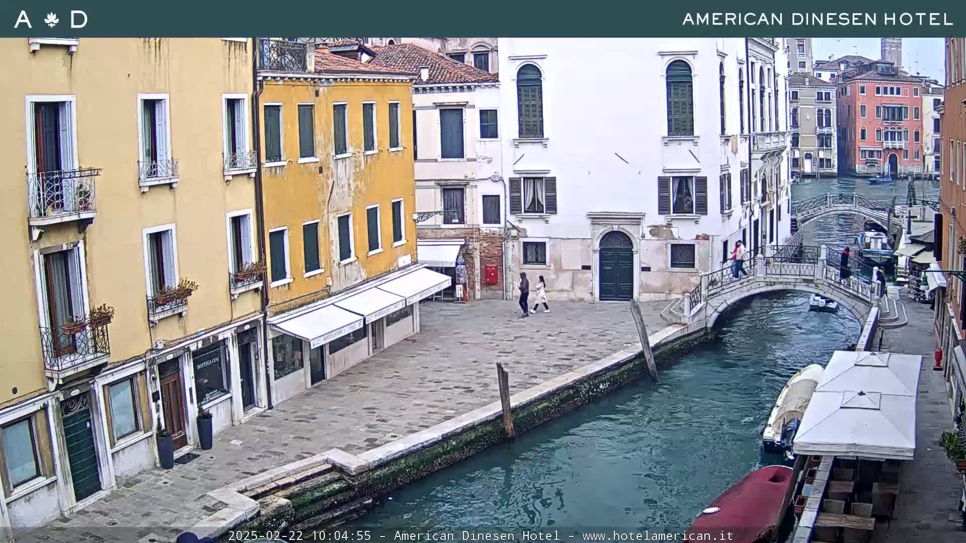 A view of a canal in Venice, Italy, shows several buildings lining the waterway, a small footbridge, and a few people walking along the canal.