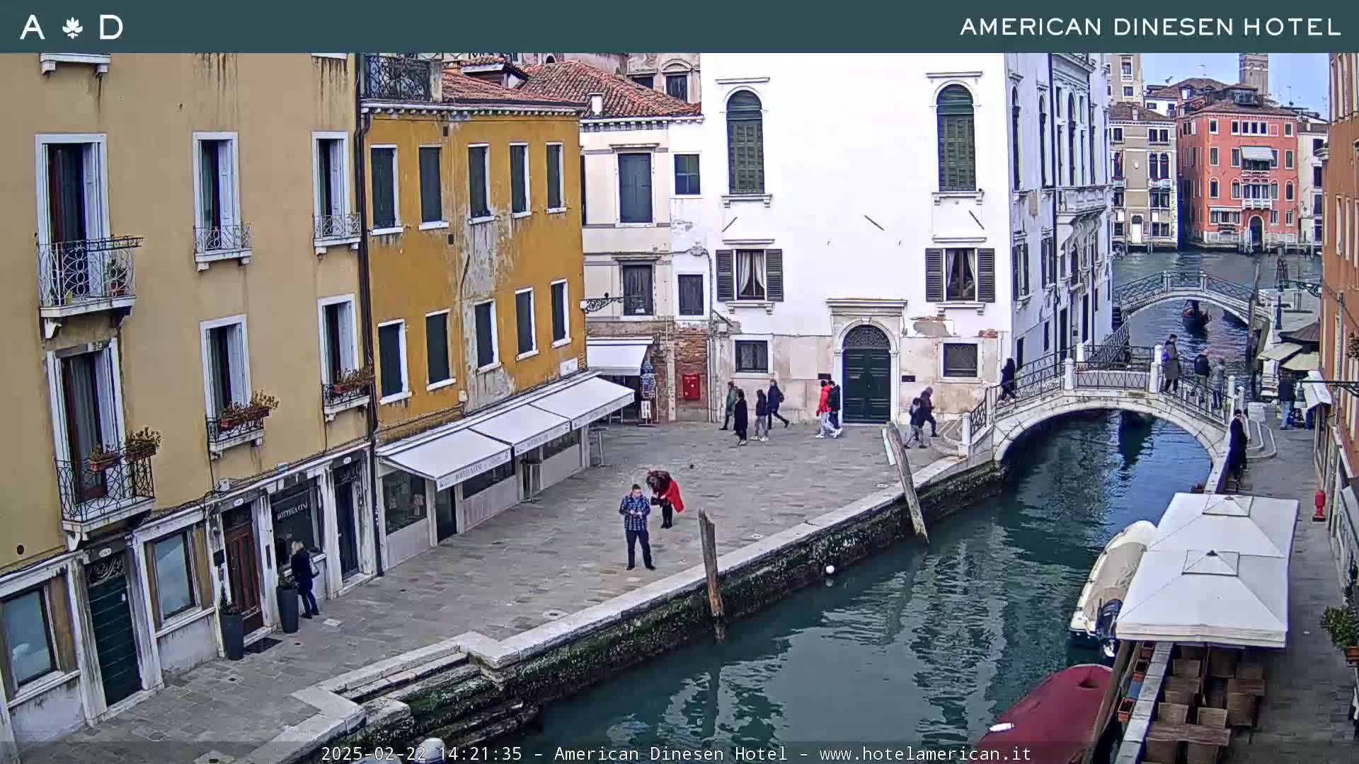 A cloudy day in Venice shows a canal with a bridge, several buildings lining the waterway, and a few people walking along the canal banks.