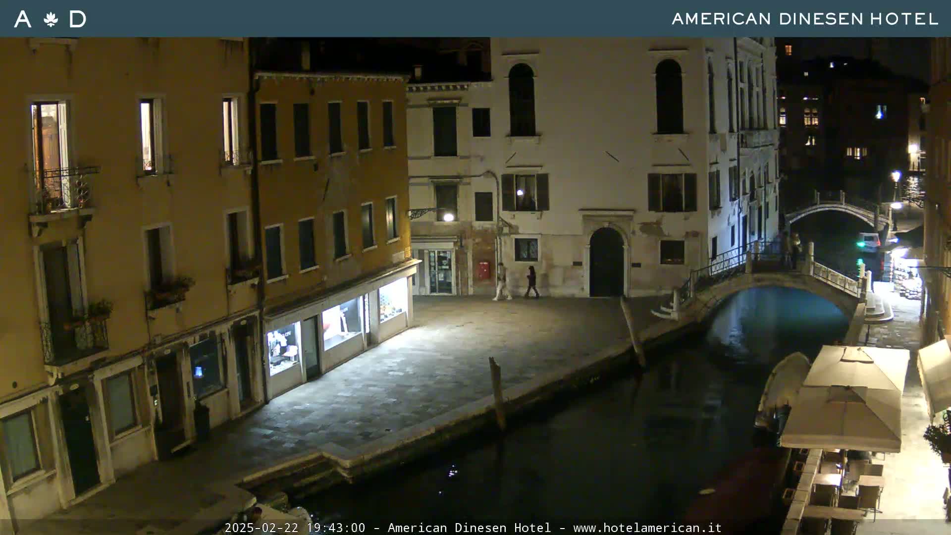 A nighttime view of a canal in Venice, Italy, shows several buildings lining the waterway, with a small bridge and two pedestrians visible.