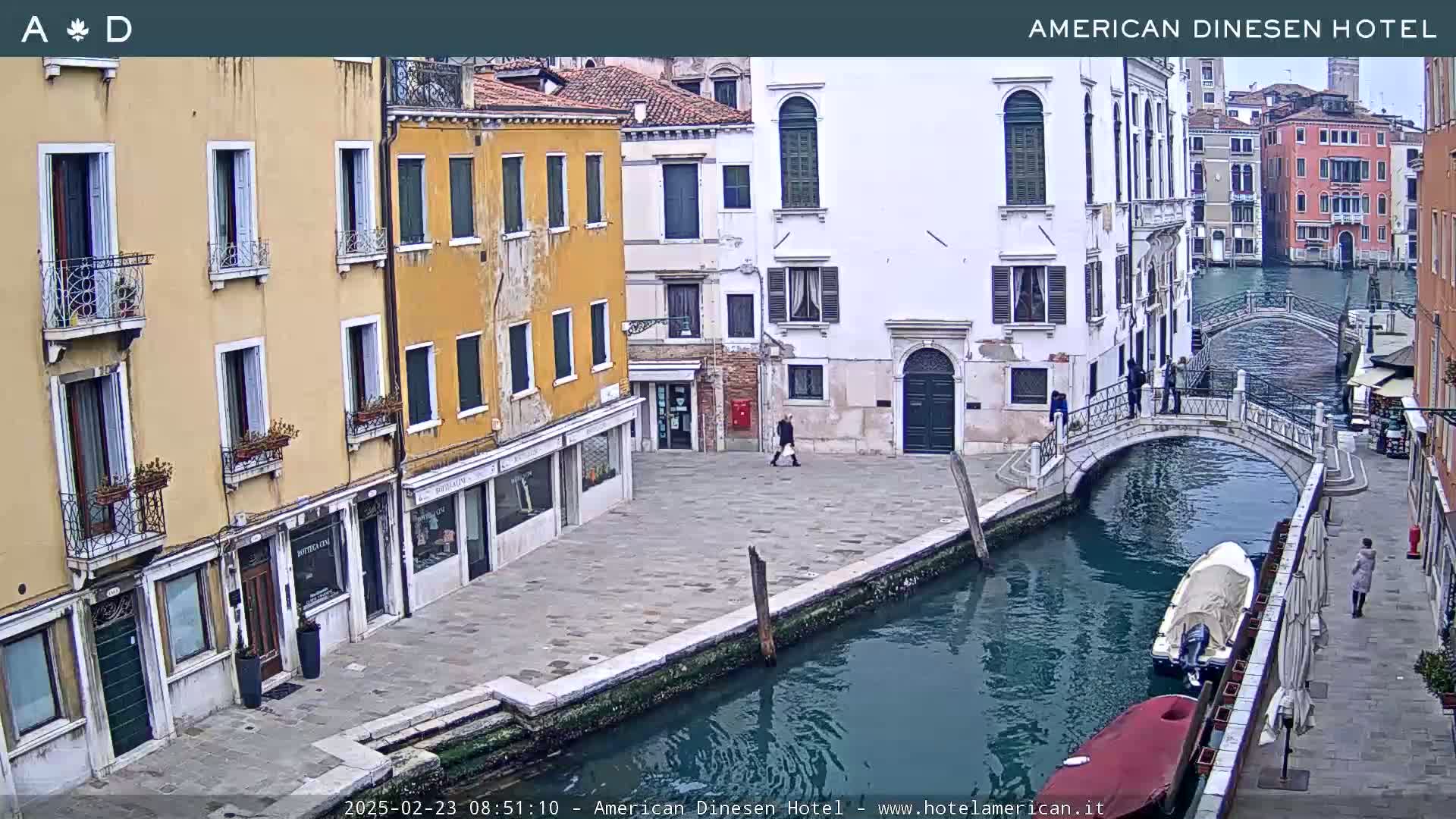A canal in Venice, Italy, with several buildings lining its banks, a small bridge crossing it, and a few pedestrians and boats visible on an overcast day.