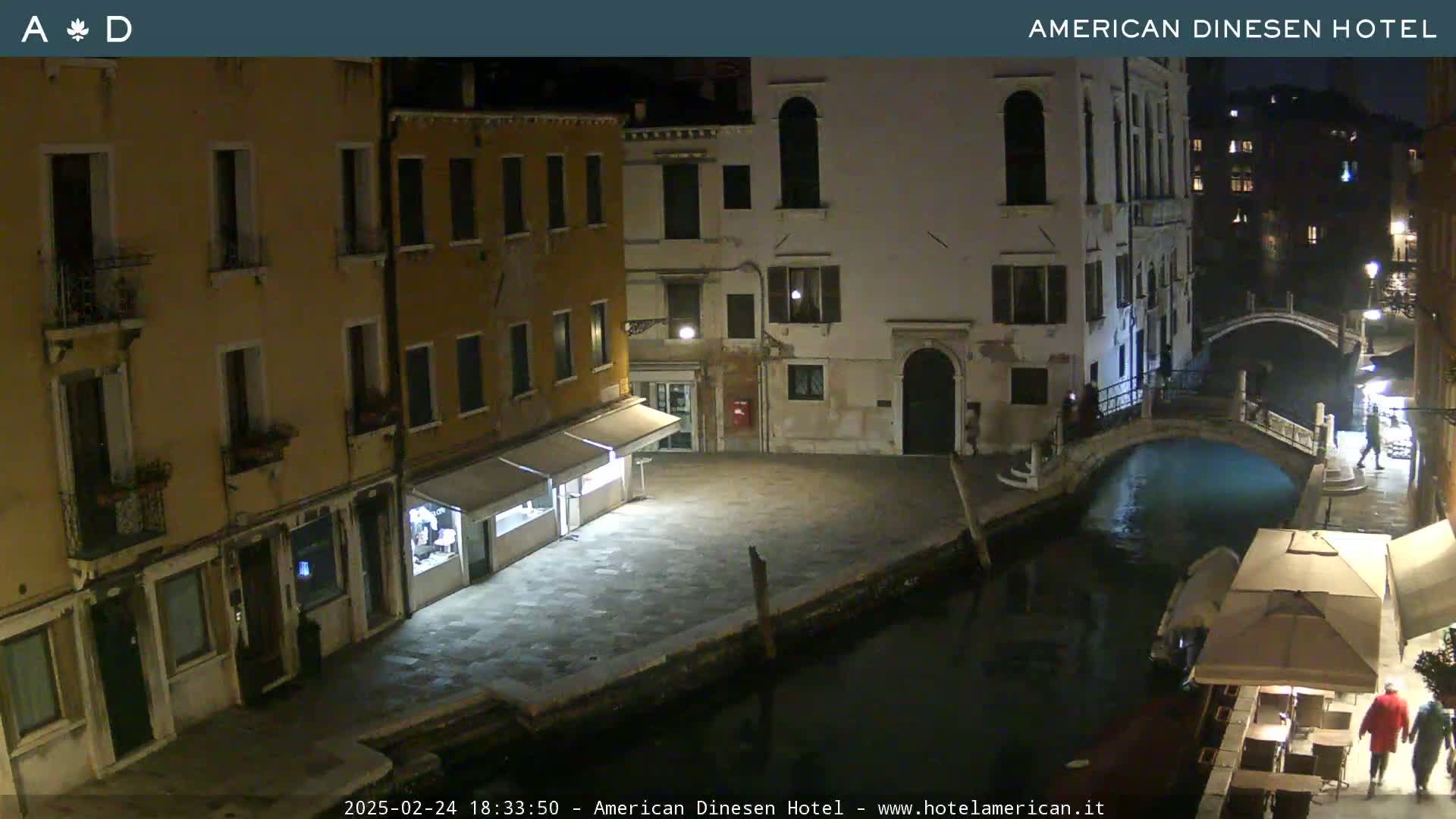 A nighttime view of a canal in Venice, Italy, shows several buildings lining the waterway, with some outdoor seating along the canal.