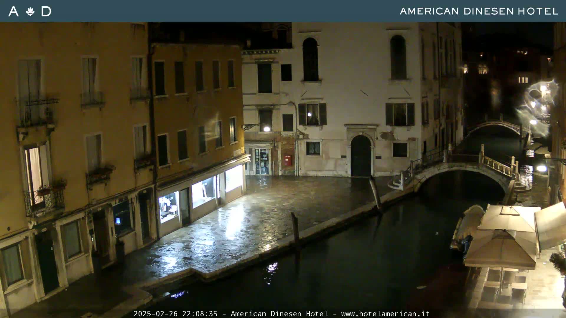 A nighttime view of a canal in Venice, showing several buildings along the waterway, a bridge, and wet cobblestones.