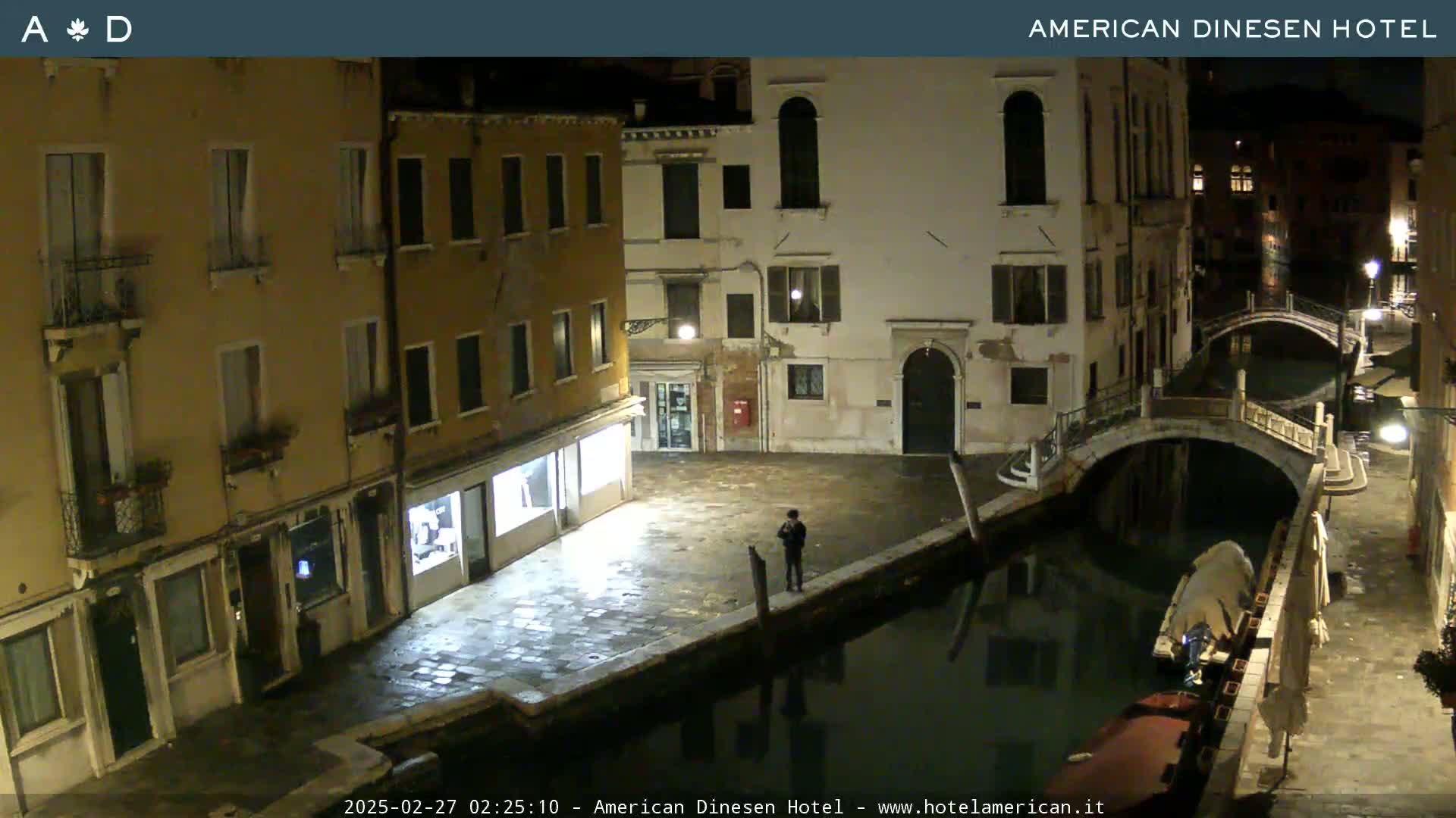 A nighttime view of a canal in Venice, showing several buildings and a small bridge, with a single person standing on a waterfront.
