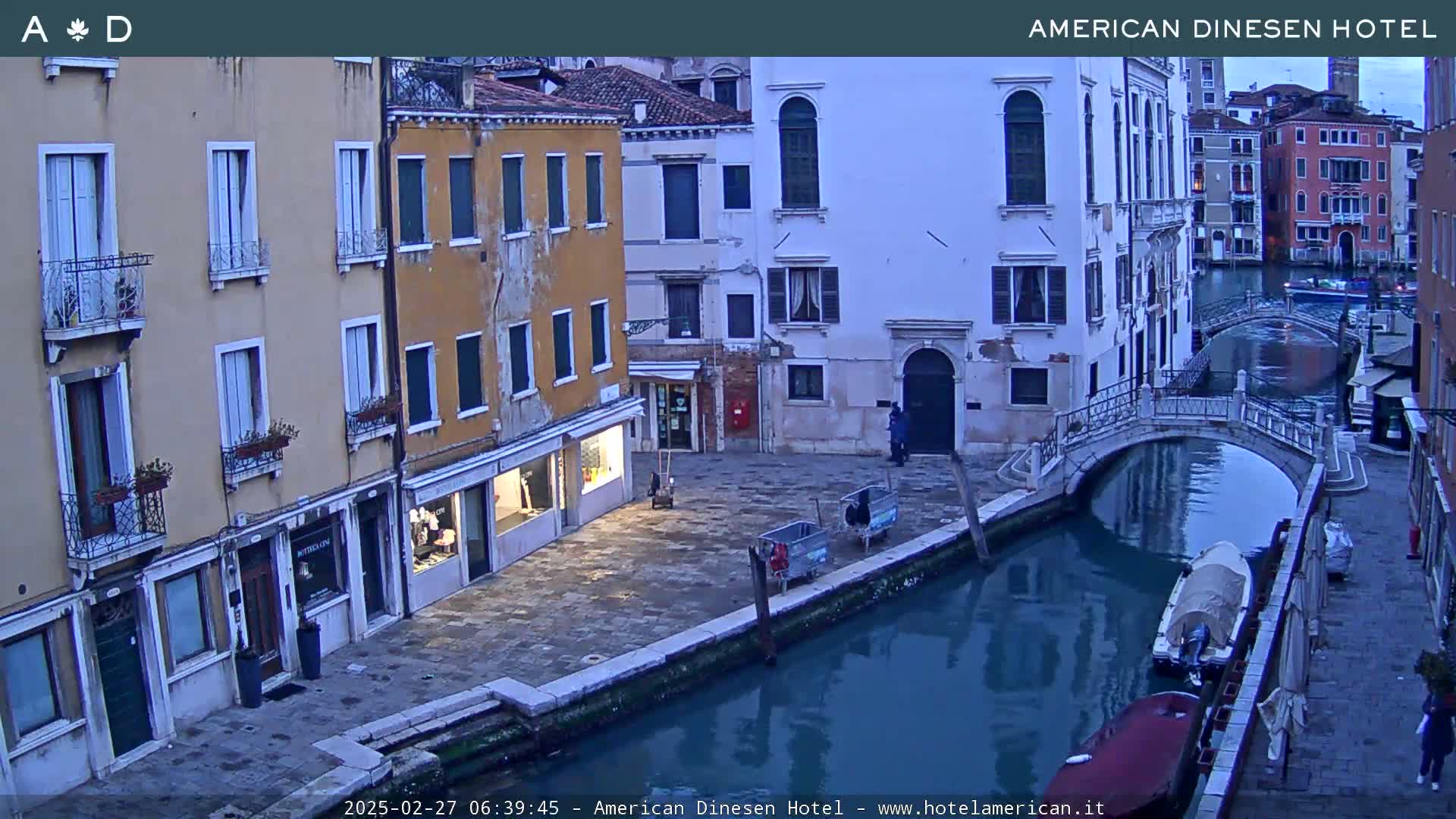 A tranquil canal scene in Venice, Italy, at dusk, shows several buildings lining the waterway, a small bridge, and a couple of boats moored along the canal.