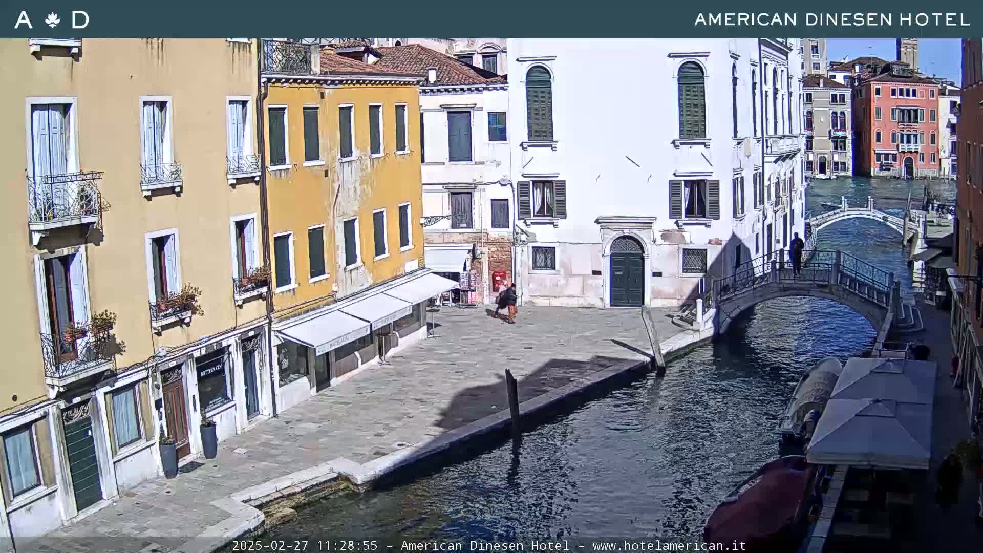A sunny view of a canal in Venice, with several buildings lining the waterway, a pedestrian bridge, and a few people walking.