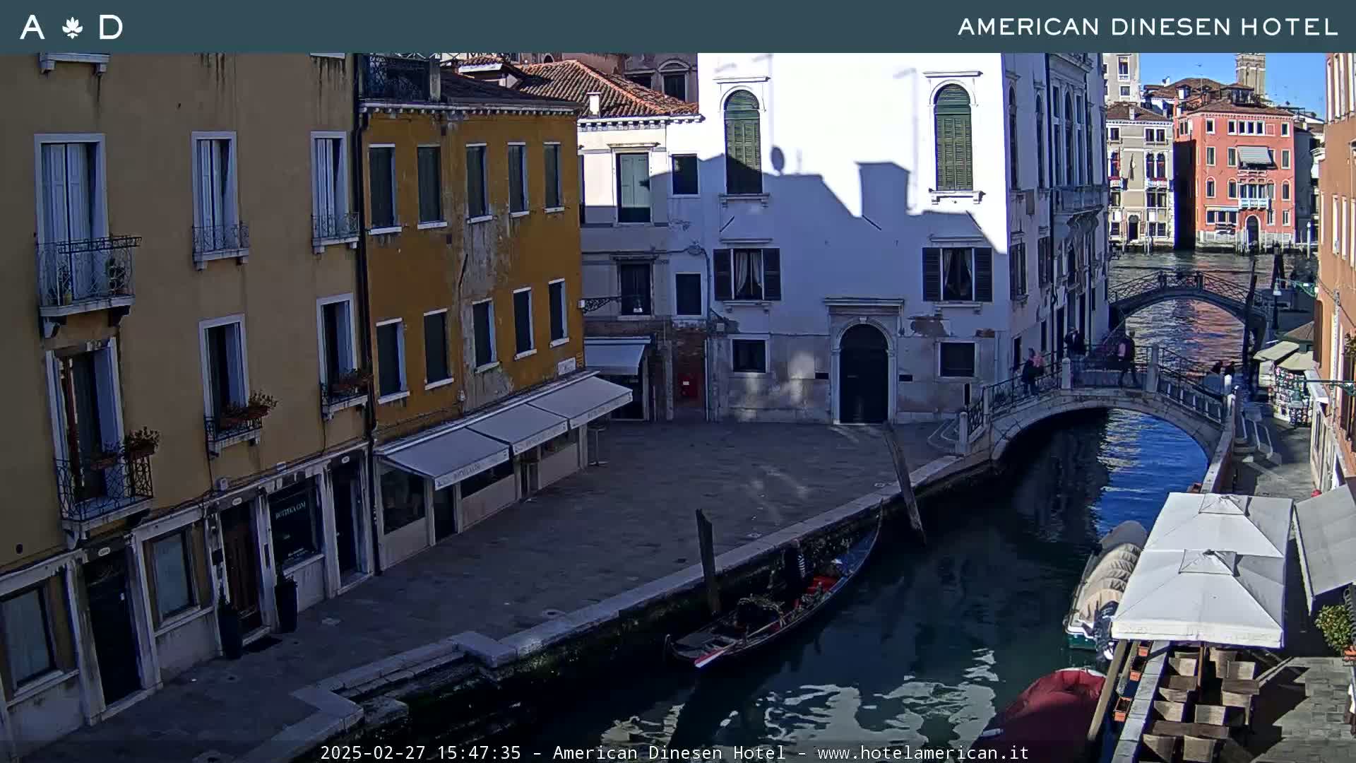 A sunny Venetian canal scene shows colorful buildings lining the waterway, a gondola moored nearby, and a small bridge with pedestrians crossing.