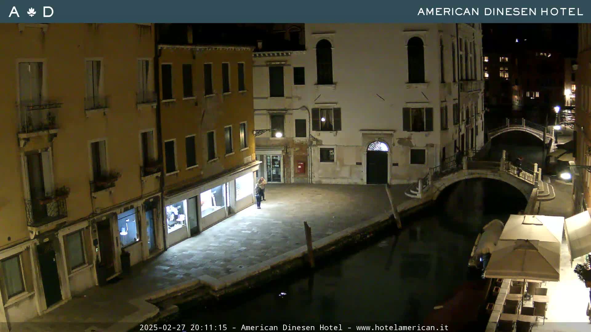 A nighttime view of a canal in Venice, showing several buildings along its banks, a small bridge, and a couple of people standing near a storefront.