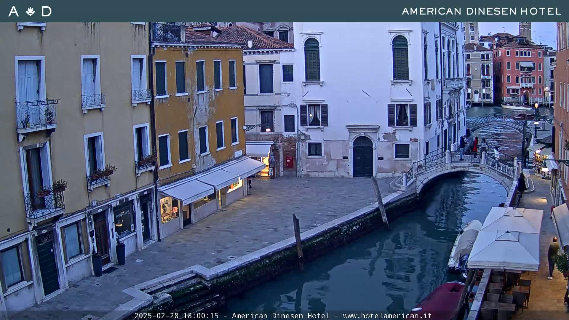 A canal in Venice, Italy, at dusk, shows several buildings along its banks, a small footbridge, and a few people walking along the water's edge.