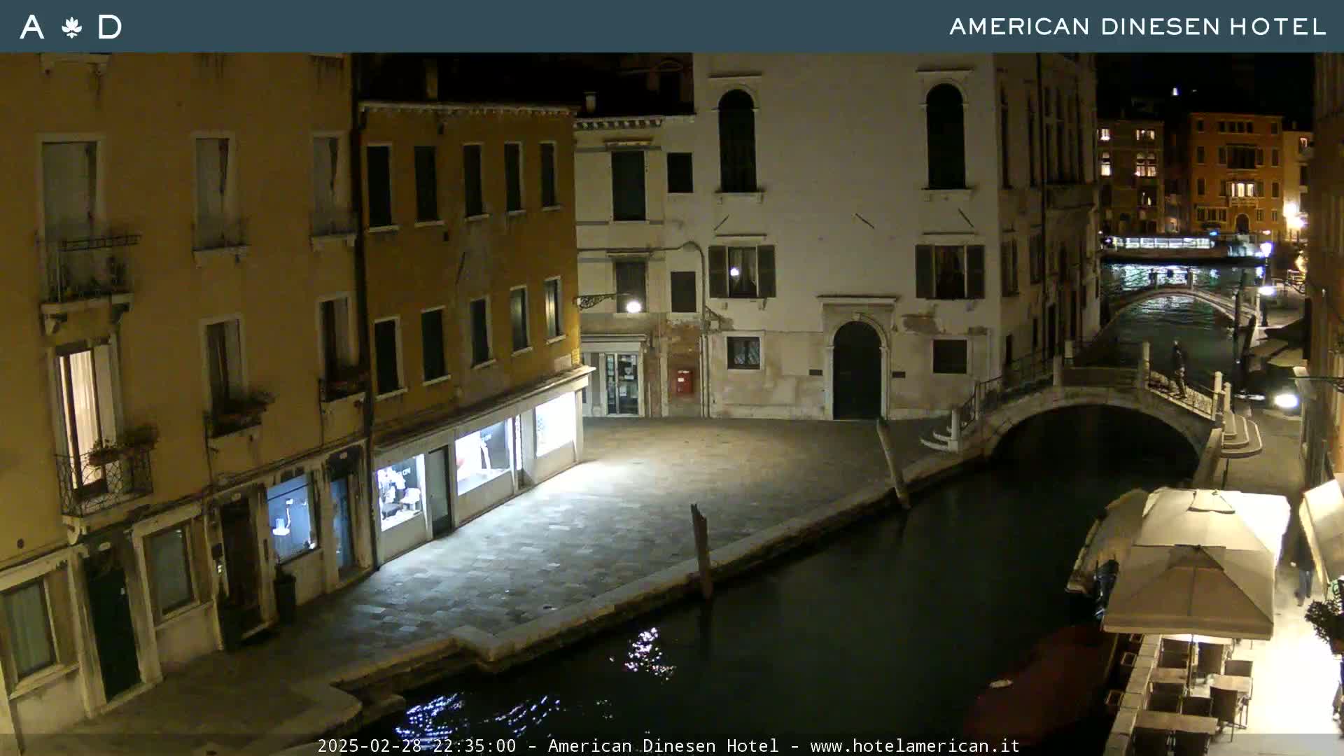 A nighttime view of a canal in Venice, showing several buildings along the waterway, a small bridge, and a patio area with outdoor seating.