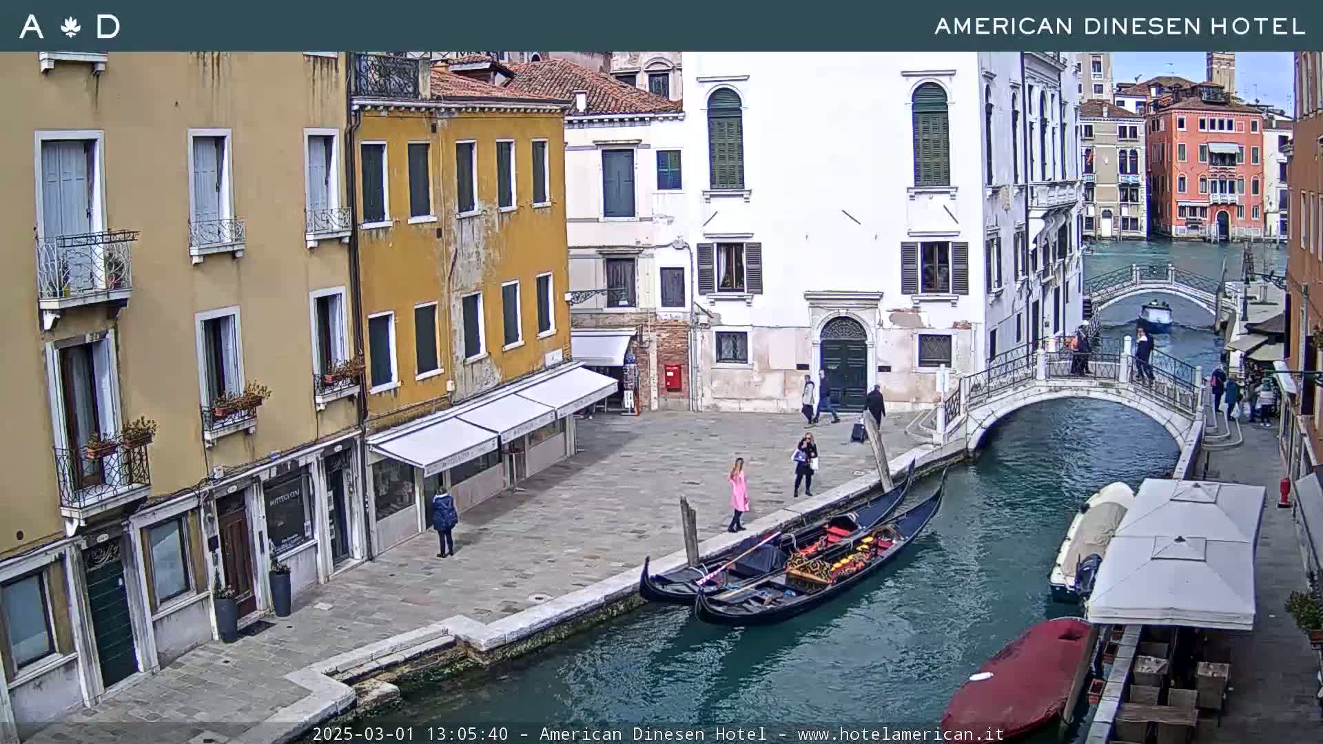A sunny view of a canal in Venice, Italy, shows several gondolas moored along a quayside lined with buildings, a small bridge, and pedestrians.