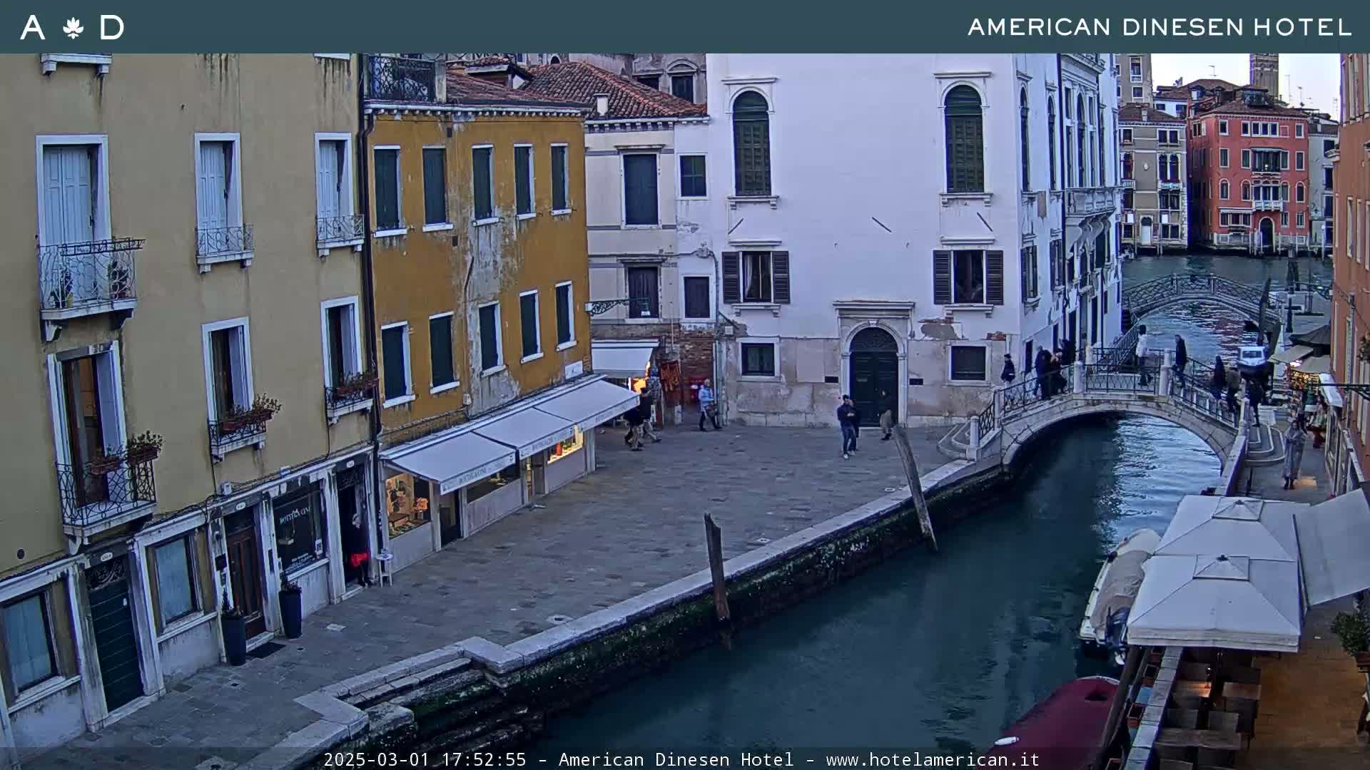 A canal in Venice, Italy, under a twilight sky, shows several buildings, a footbridge, and a few people walking along the waterfront.