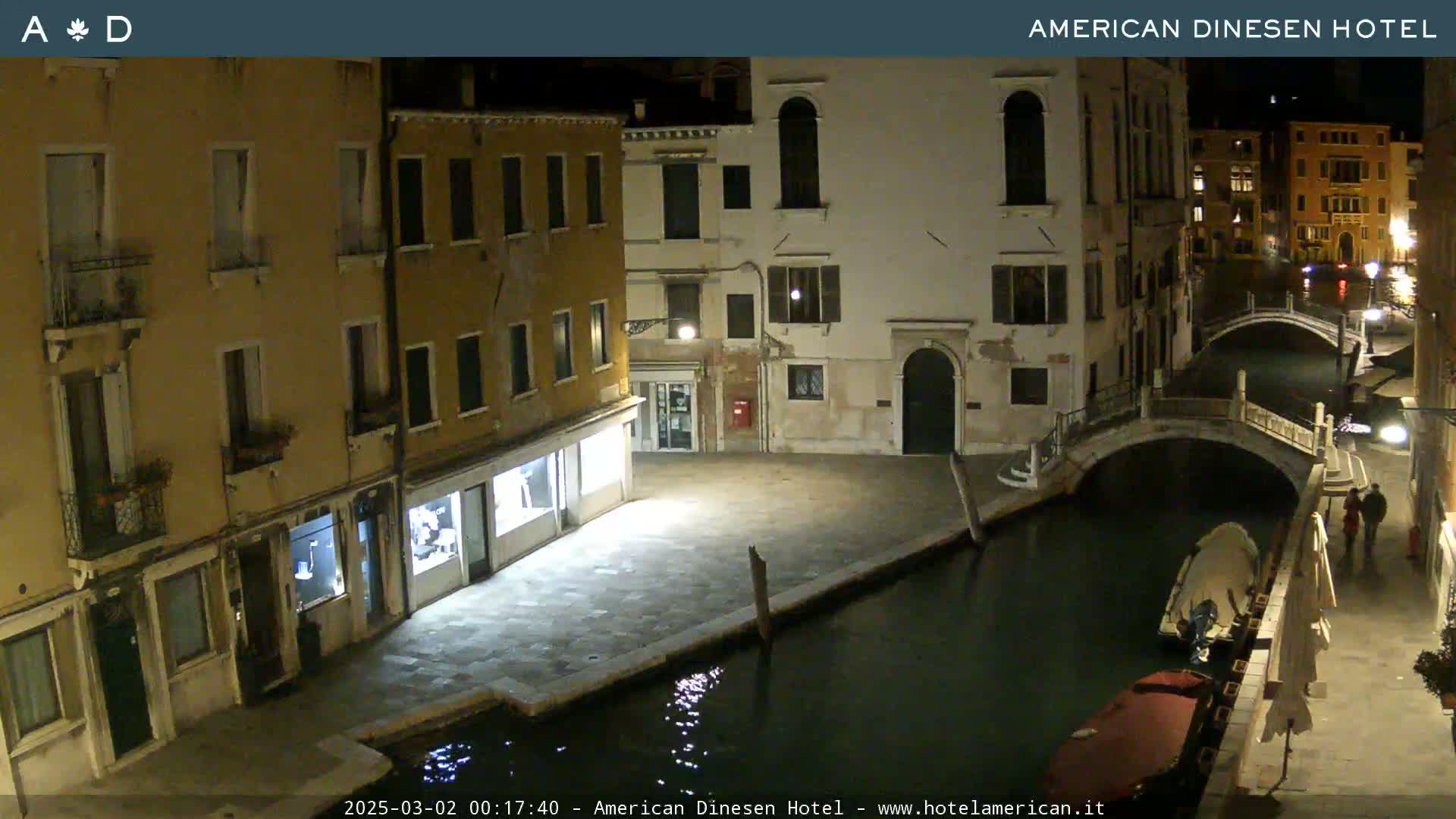 A nighttime view of a canal in Venice, showing several buildings lining the waterway, a small bridge, and a gondola.