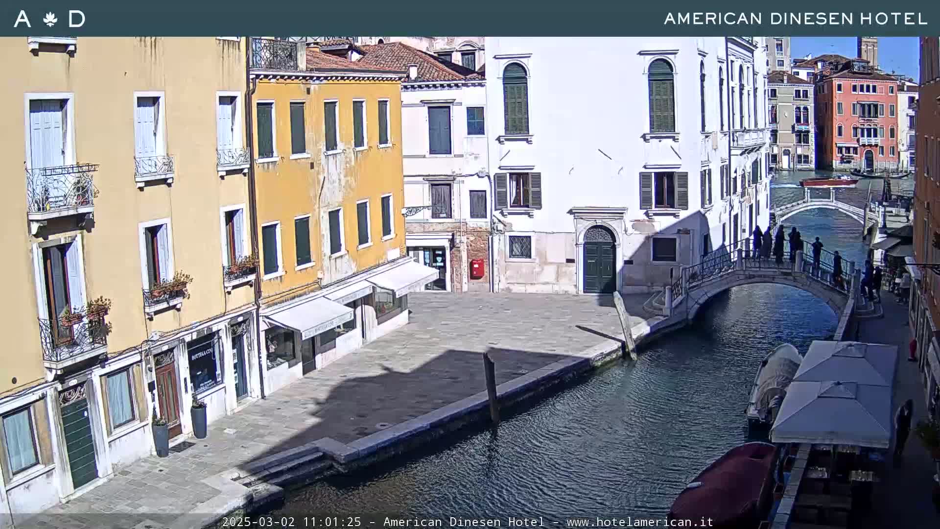 A sunny view of a canal in Venice, Italy, shows colorful buildings lining the waterway, a bridge with pedestrians, and a few boats.