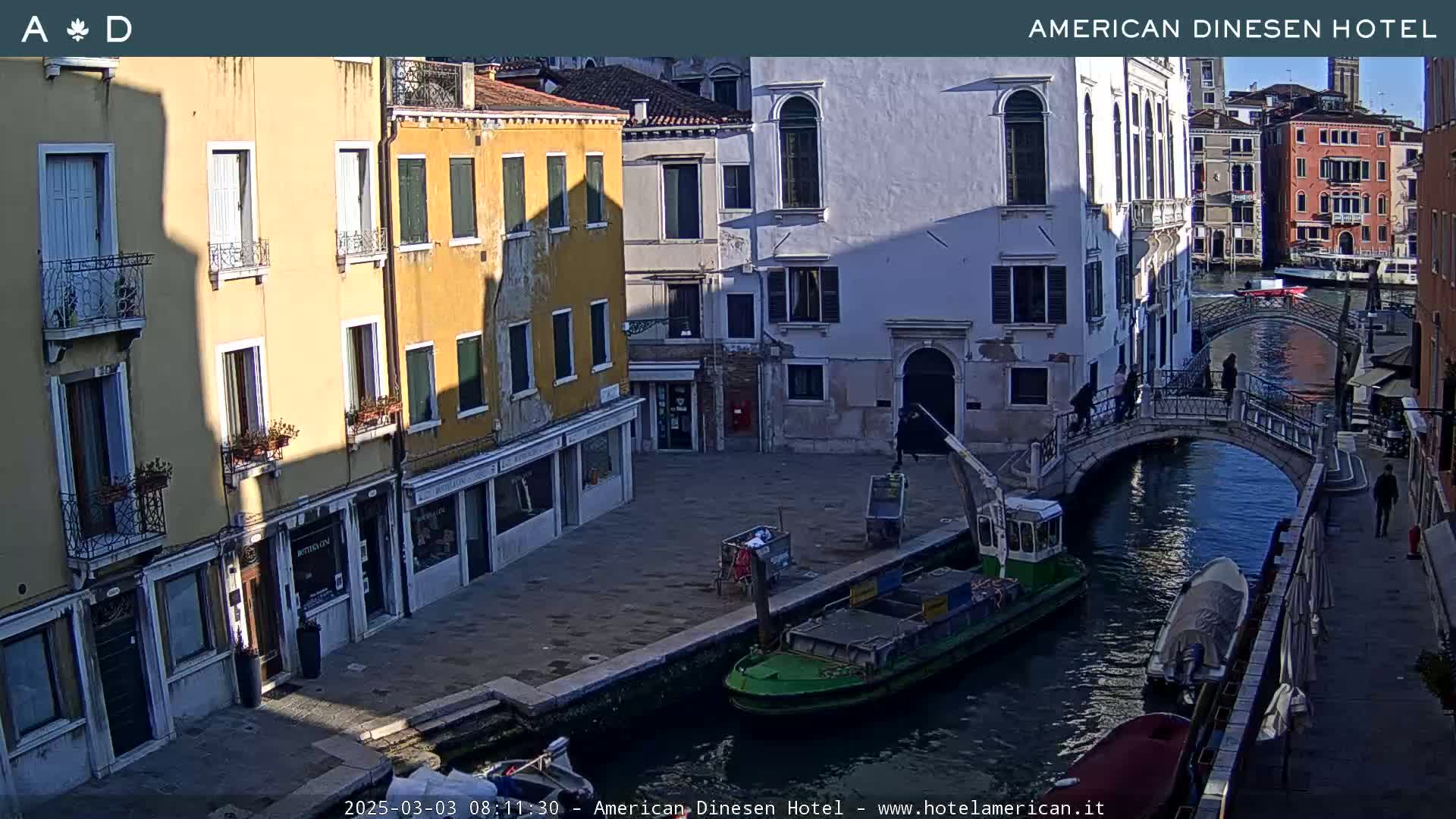 A sunny day in Venice shows a canal with a small bridge, boats, and colorful buildings lining the waterway.