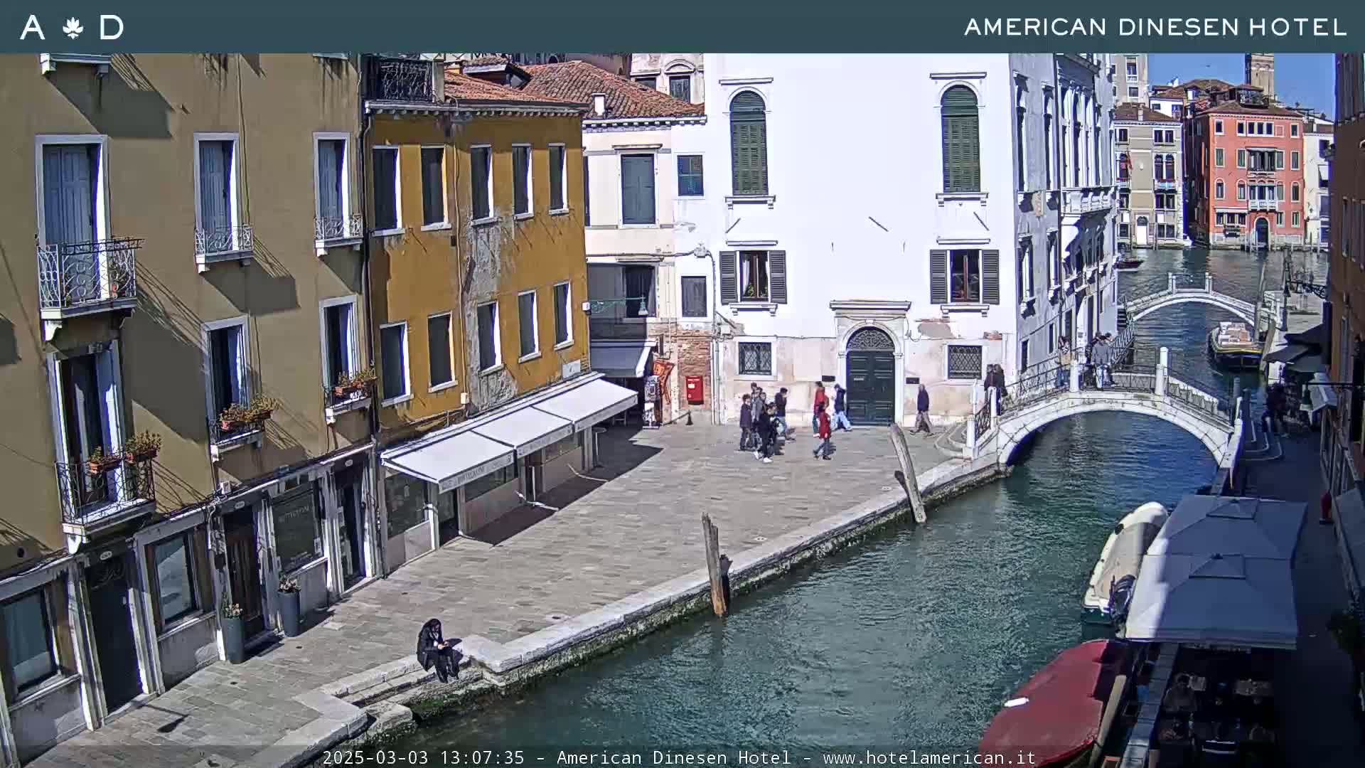 A sunny day in Venice shows a canal with a bridge, boats, and several buildings lining the waterway, with a few people walking along the canal.