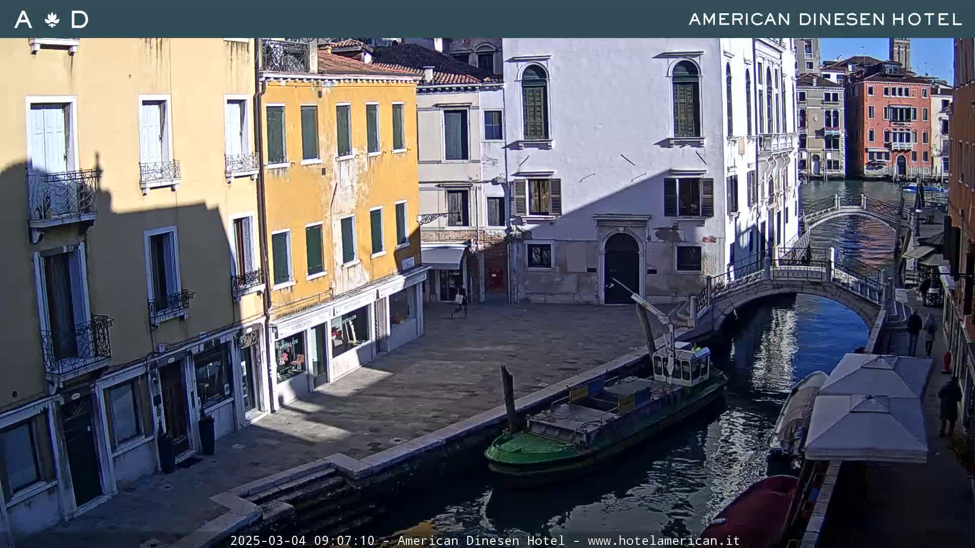 A sunny day in Venice shows a canal with a small boat, a stone bridge, and several buildings lining the waterway.