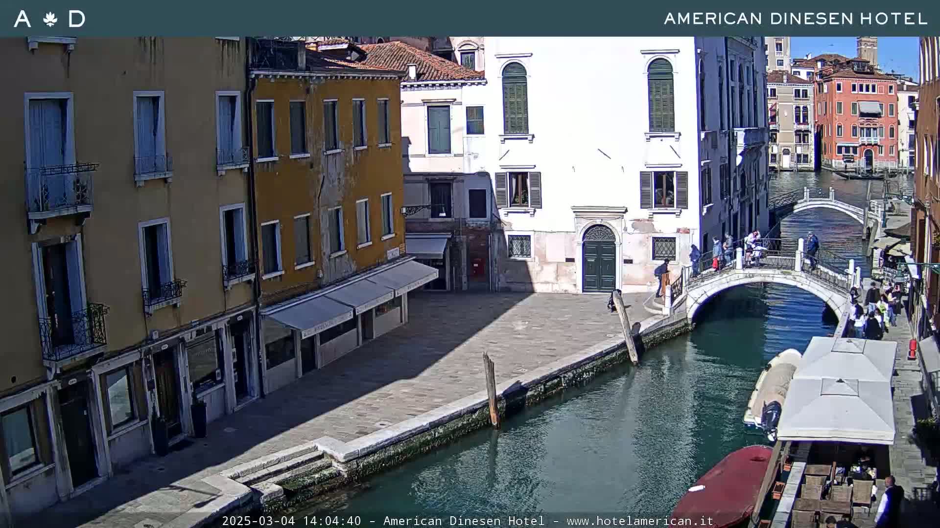 A sunny day in Venice shows a canal with a bridge, several buildings along the waterway, and a boat docked nearby.