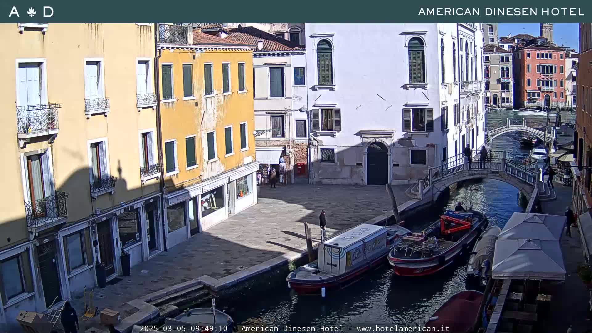 A sunny day in Venice shows several buildings lining a canal, with boats moored along the waterway and pedestrians crossing a small bridge.