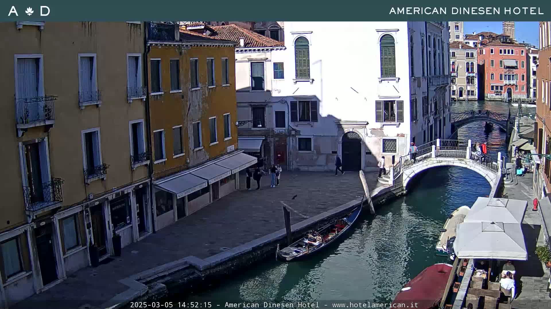 A sunny Venetian canal scene features a gondola moored near a bridge, with several buildings lining the waterway and people walking along the canal side.