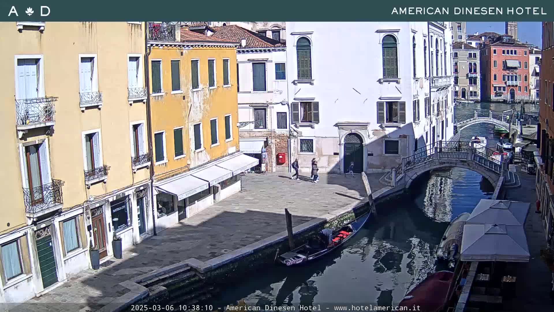 A sunny day in Venice shows a canal with a gondola, a small bridge, and colorful buildings lining the waterway.
