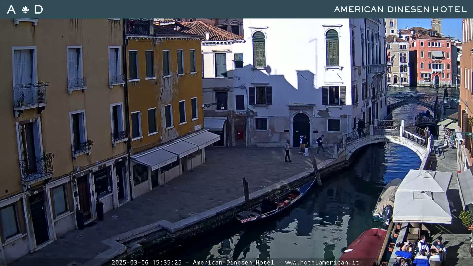A sunny view of a canal in Venice, Italy, shows several buildings, a gondola, a small bridge, and some people walking along the canal.