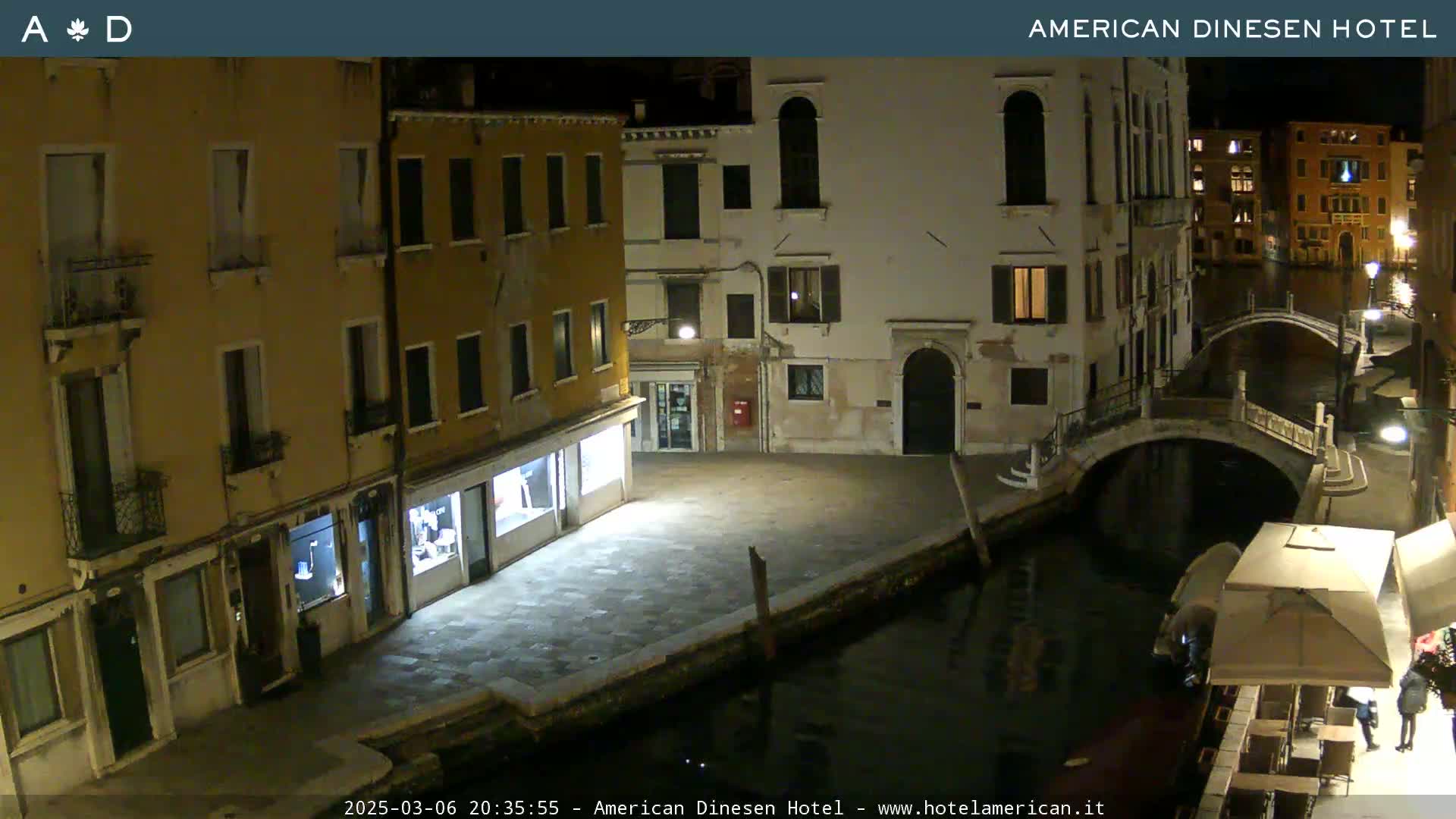 A nighttime view of a canal in Venice, Italy, shows several buildings lining the waterway, with a small bridge visible in the distance.