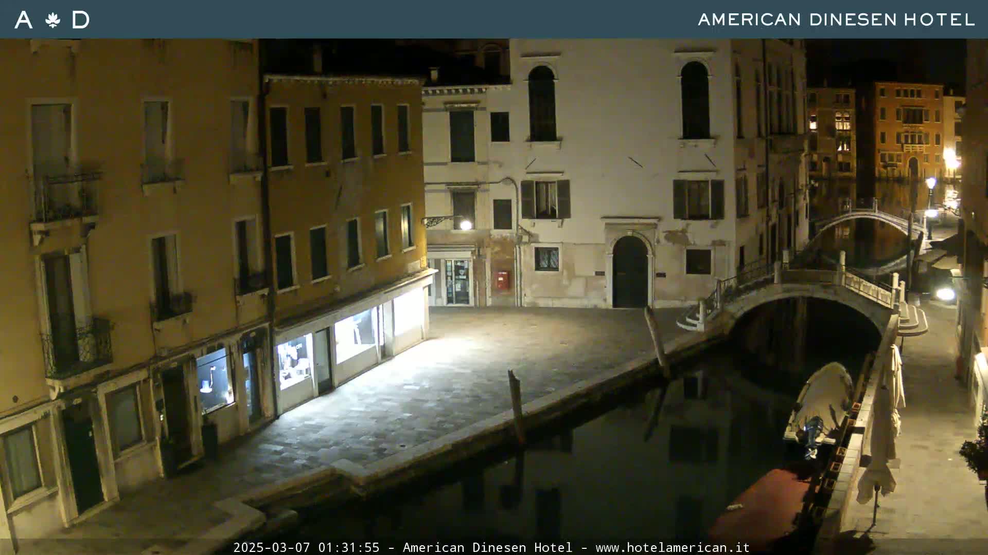 A nighttime view of a canal in Venice, Italy, shows several buildings lining the waterway and a small bridge crossing the canal; a boat is moored on the canal's edge.