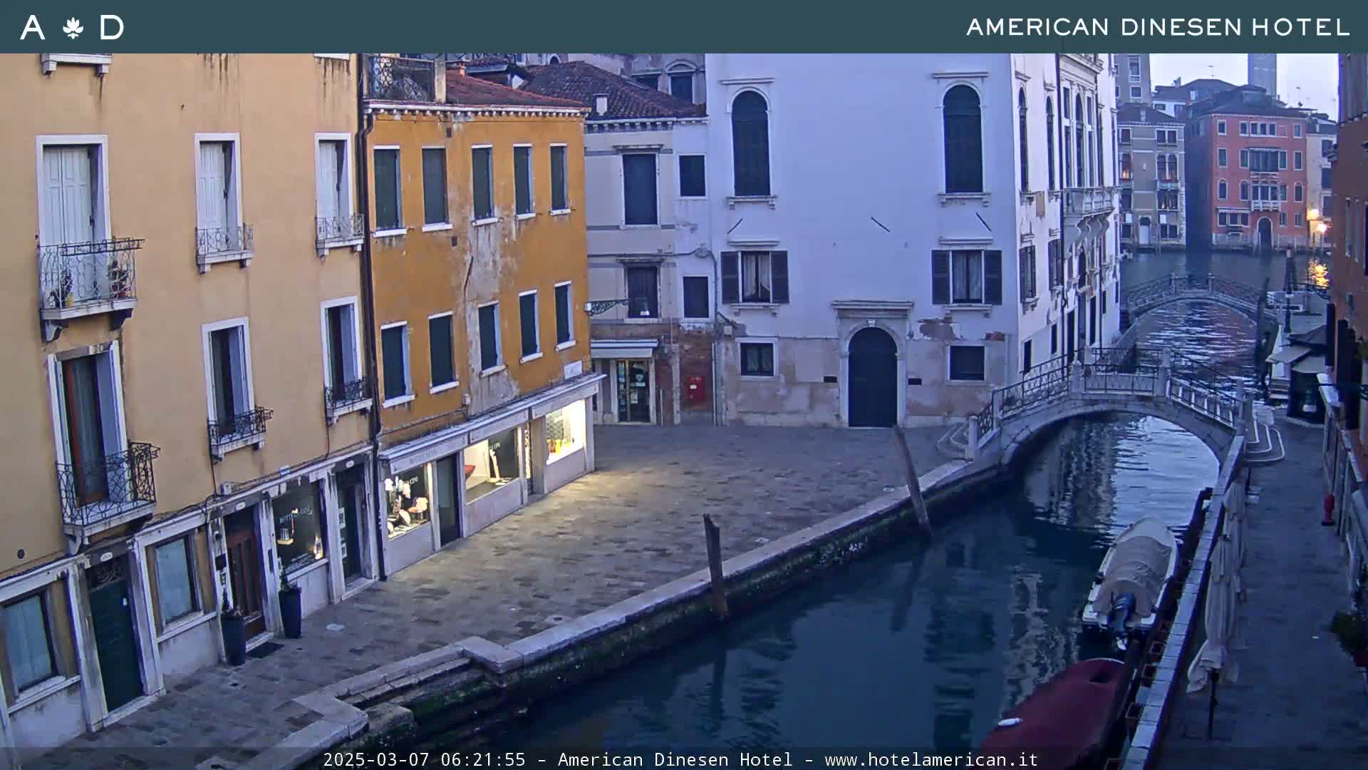 A tranquil canal scene in Venice shows several buildings lining the waterway, a small bridge spanning the canal, and a couple of boats moored alongside the buildings under a dusky sky.