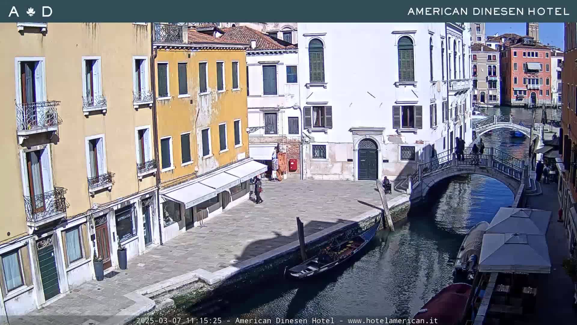 A sunny day in Venice shows a canal with gondolas, a pedestrian bridge, and several buildings along the waterway.