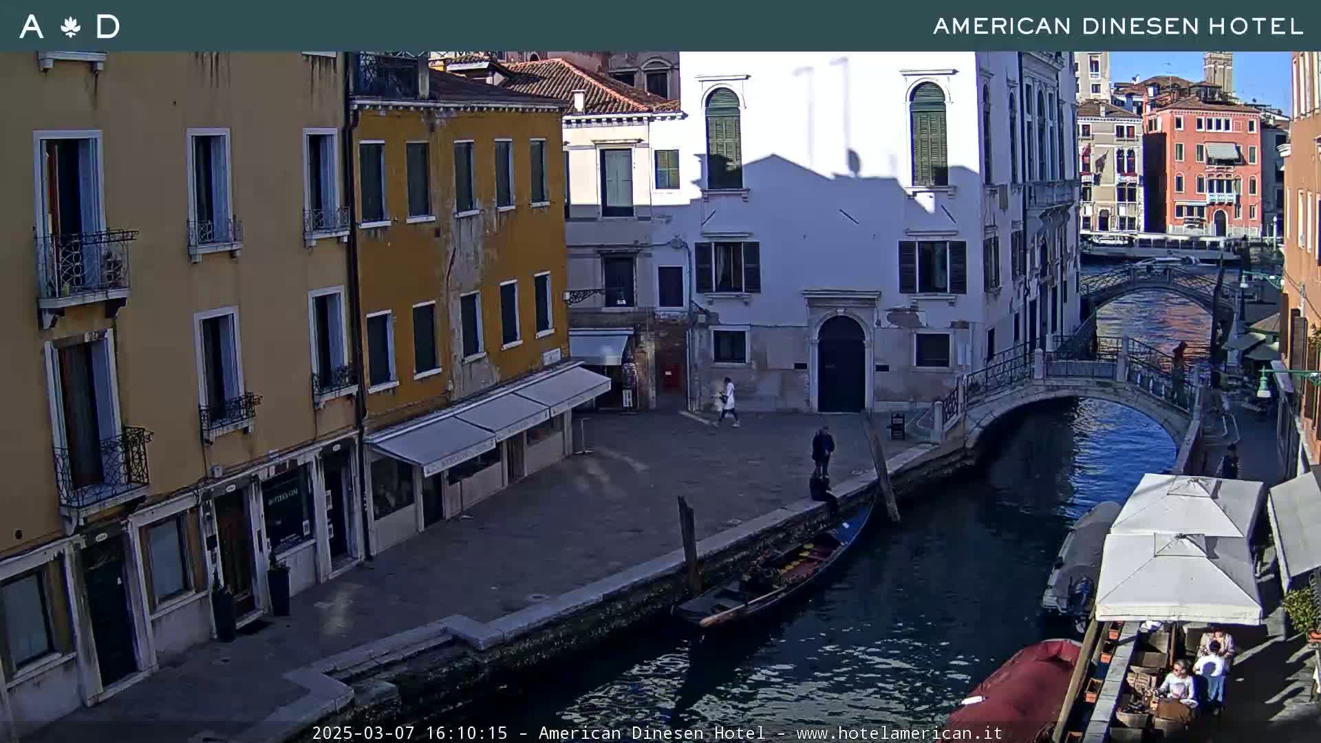 A sunny Venetian canal scene shows several buildings lining the waterway, a gondola moored nearby, and a small arched bridge crossing the canal.