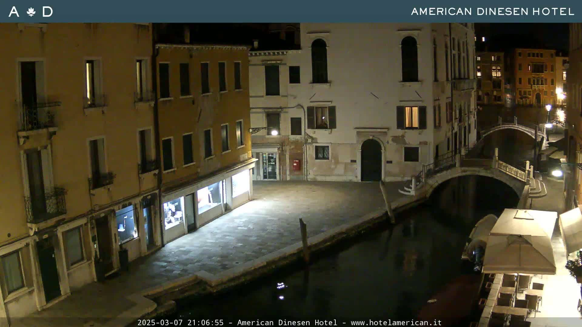 A nighttime view of a canal in Venice, showing buildings with lights on, a stone bridge, and a few outdoor cafe chairs and umbrellas.