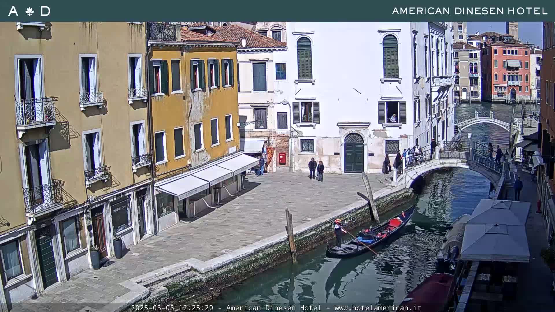 A sunny Venetian canal scene shows a gondola on the water, pedestrians on a stone bridge and along the canal-side walkways, and several buildings lining the waterway.