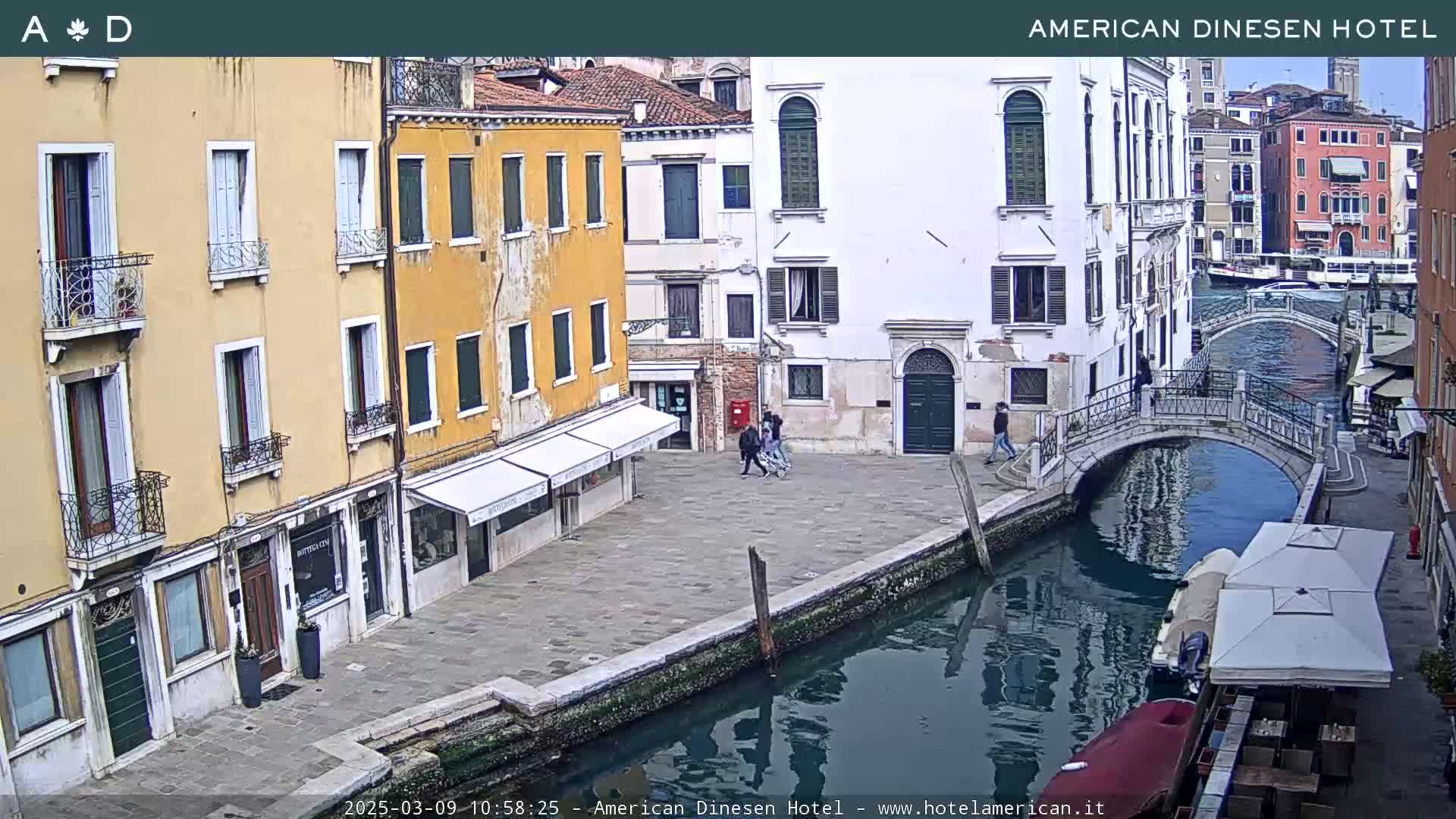 A canal in Venice, Italy, with several buildings lining its banks, a small bridge crossing it, and a few pedestrians visible on a partly sunny day.
