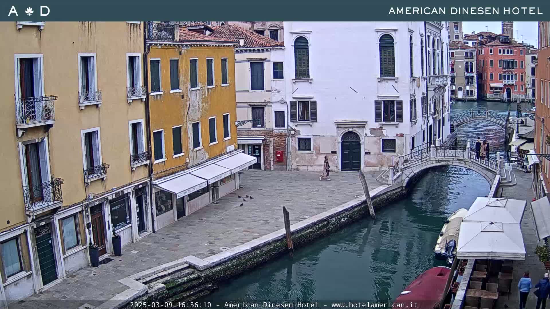A view of a canal in Venice, Italy, on an overcast day, shows several buildings lining the waterway, a small footbridge, and a few people walking along the canal.