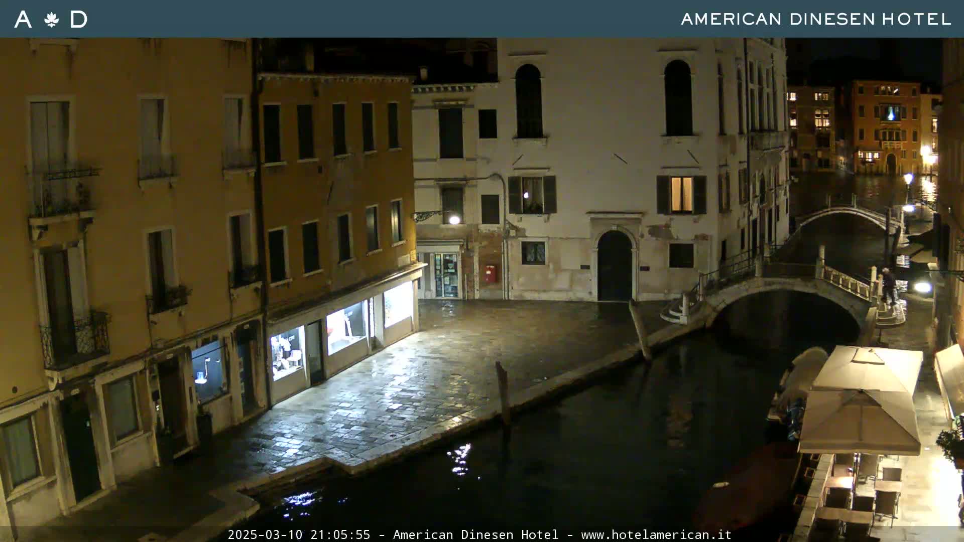 A nighttime view of a canal in Venice, showing several buildings lining the waterway, a small bridge, and a patio with outdoor seating.