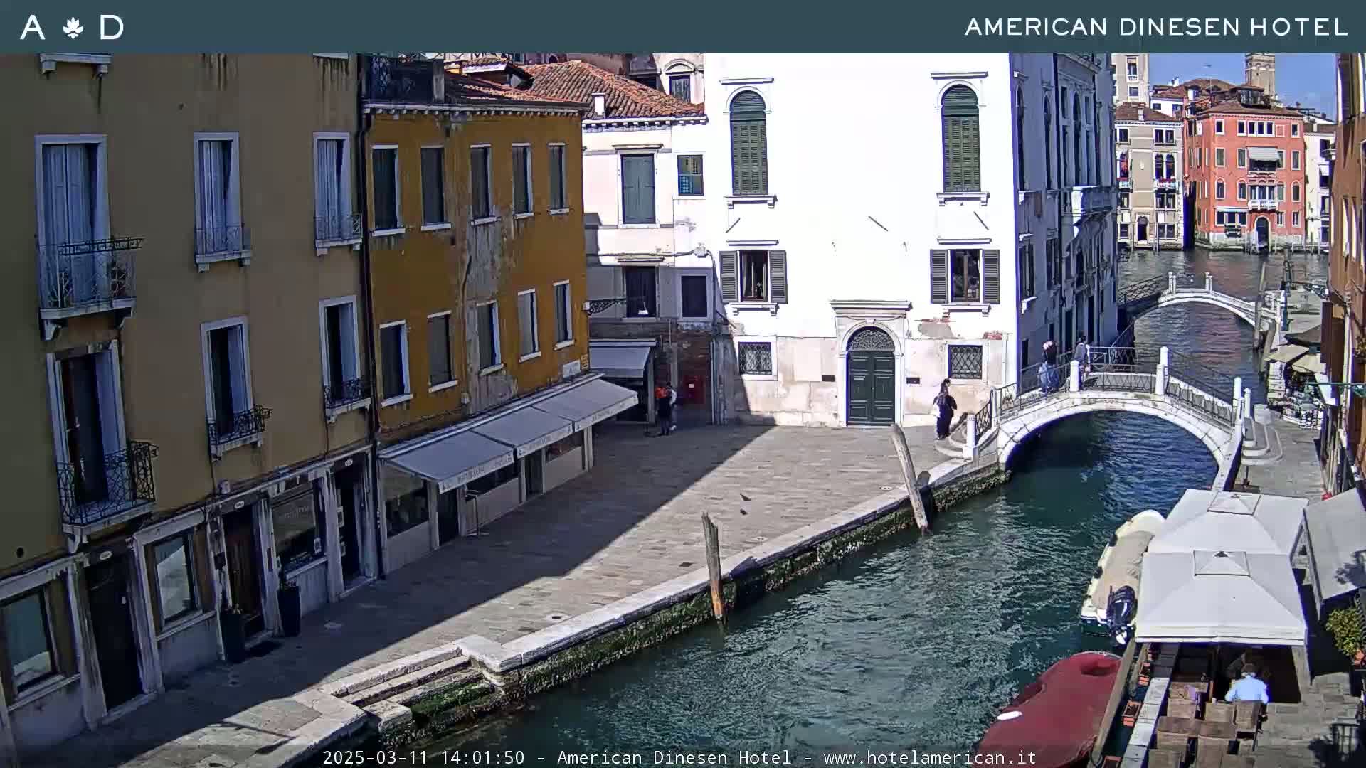 A sunny day in Venice shows a canal with a stone bridge, several buildings lining the waterway, and a small boat moored near a restaurant's outdoor seating.