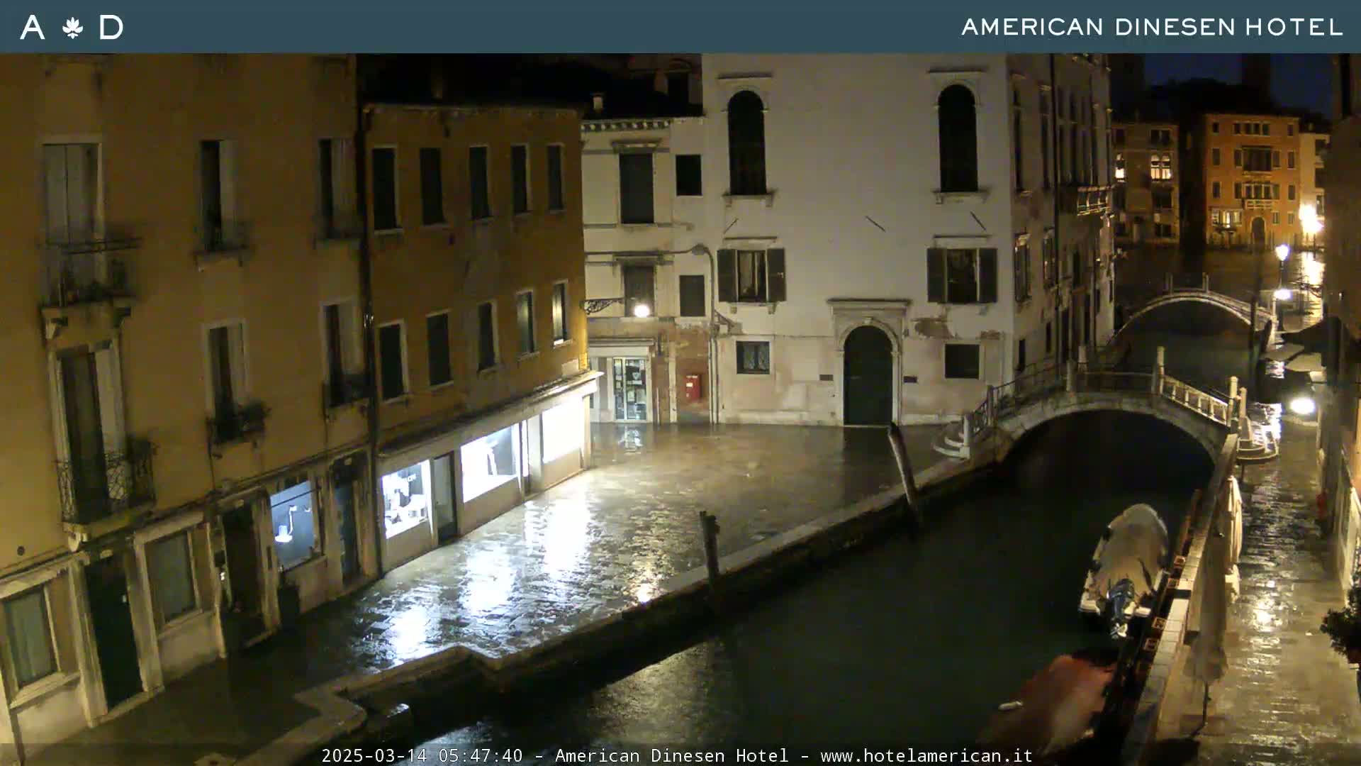 A nighttime view of a canal in Venice, Italy, shows buildings lining the waterway, a small bridge, and a boat, under rainy conditions.